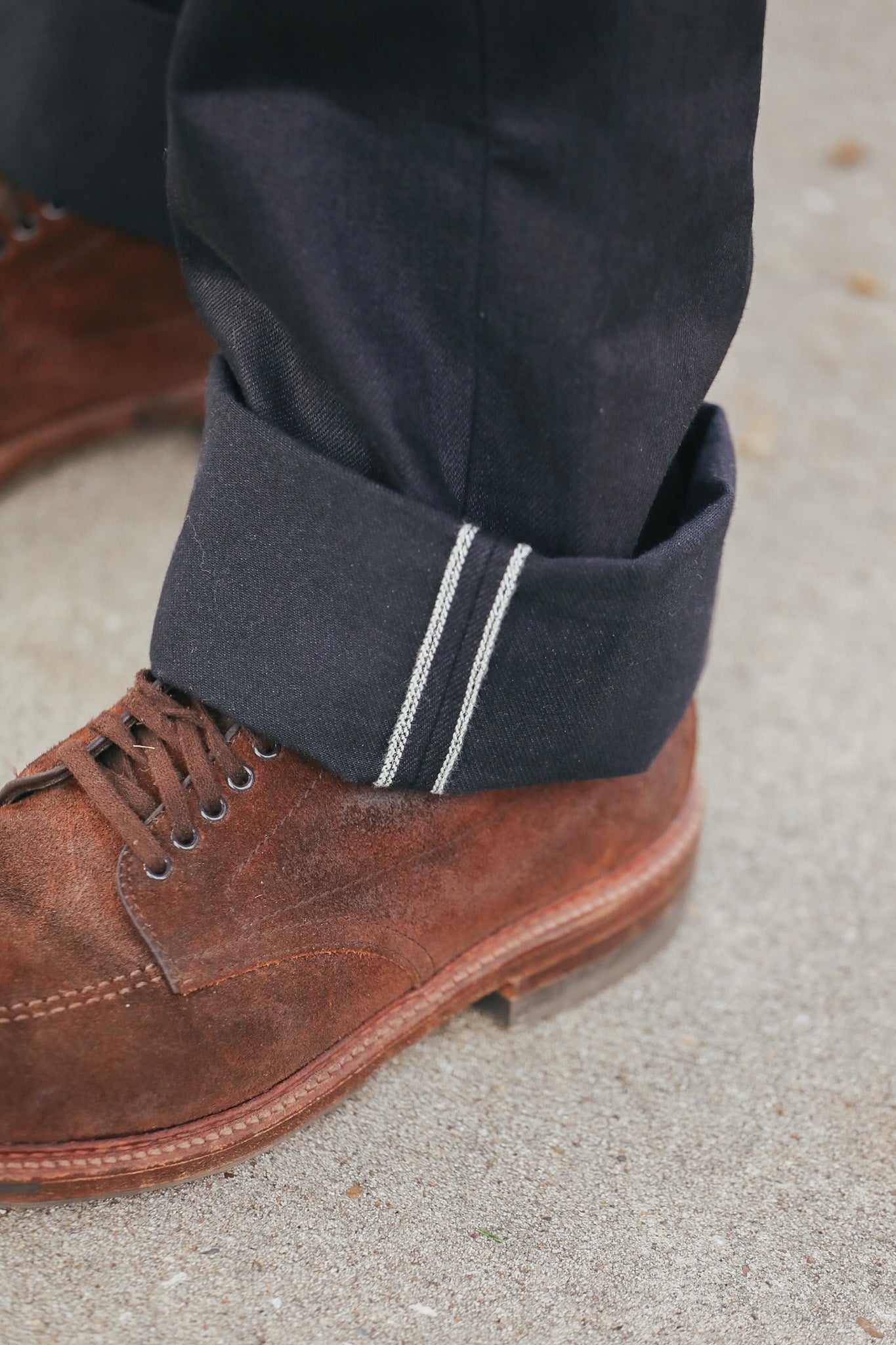 Close-up of a brown leather shoe with rolled-up black pants on a concrete surface