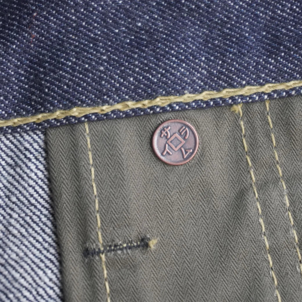 Close-up of denim and olive fabric with yellow stitching and a copper rivet button featuring Japanese characters