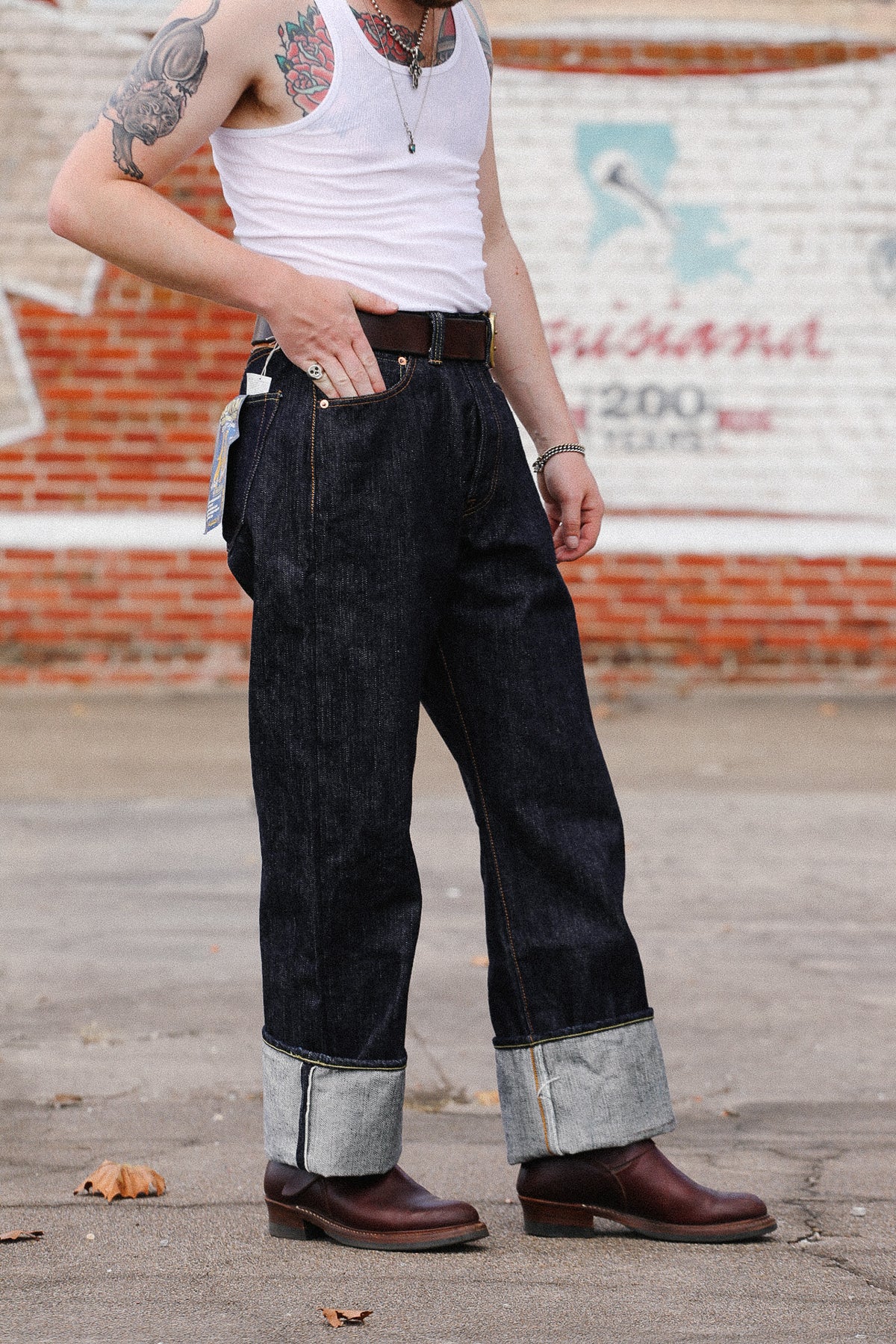 A person wearing dark indigo cuffed denim jeans, a white tank top, and brown leather boots, standing outdoors in front of a brick wall with a painted sign.
