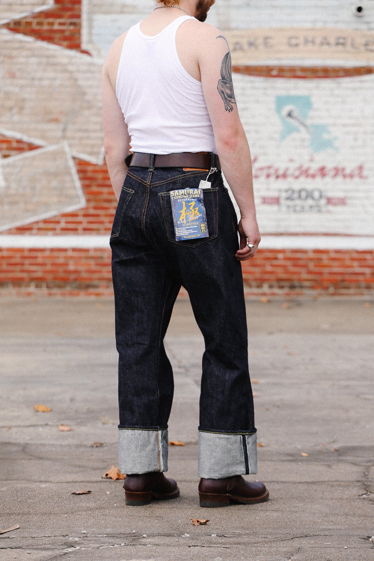 A man viewed from behind wearing dark blue cuffed denim jeans, a white tank top, and brown boots, standing outdoors in front of a brick and painted wall.