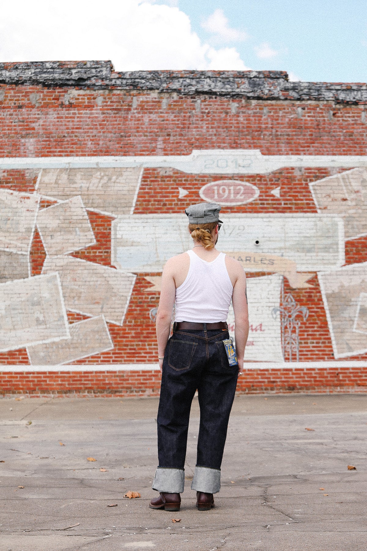 A person standing with their back to the camera in front of a brick wall with a large white painted mural, wearing a white tank top, dark blue jeans with cuffed hems, and a gray cap.