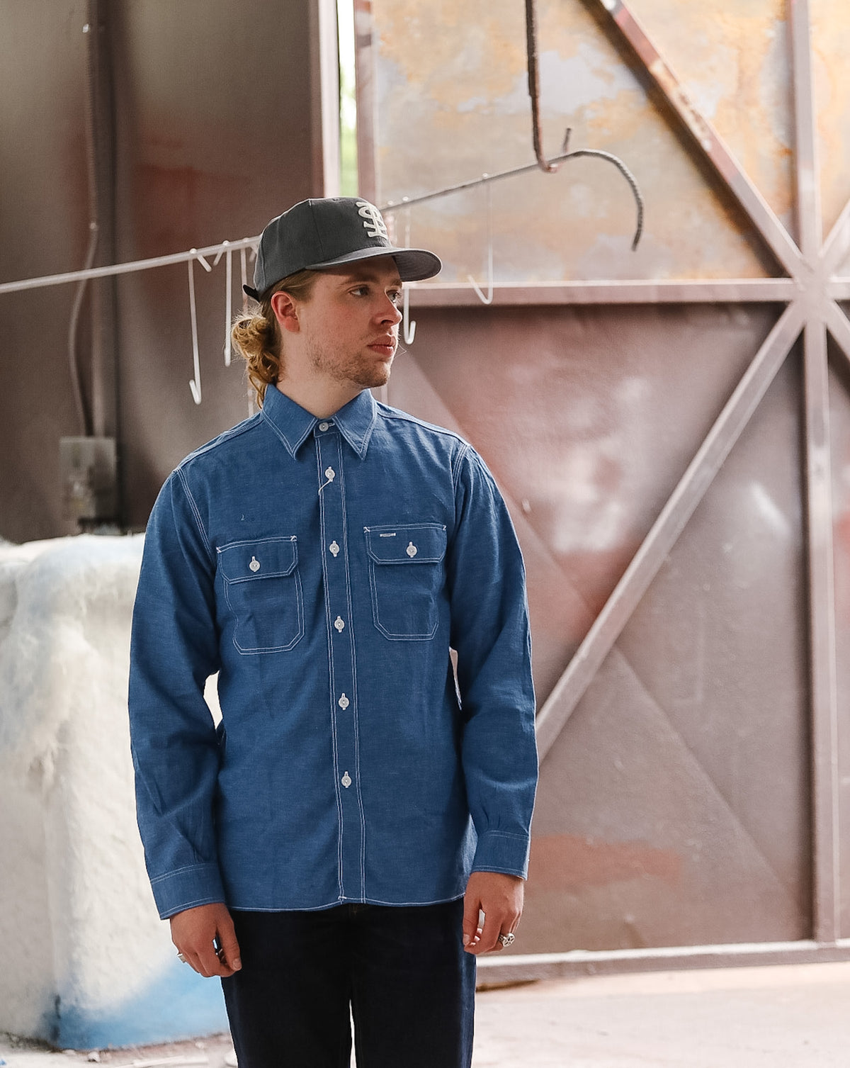 Man wearing a blue denim shirt and black cap standing in front of a textured wall.