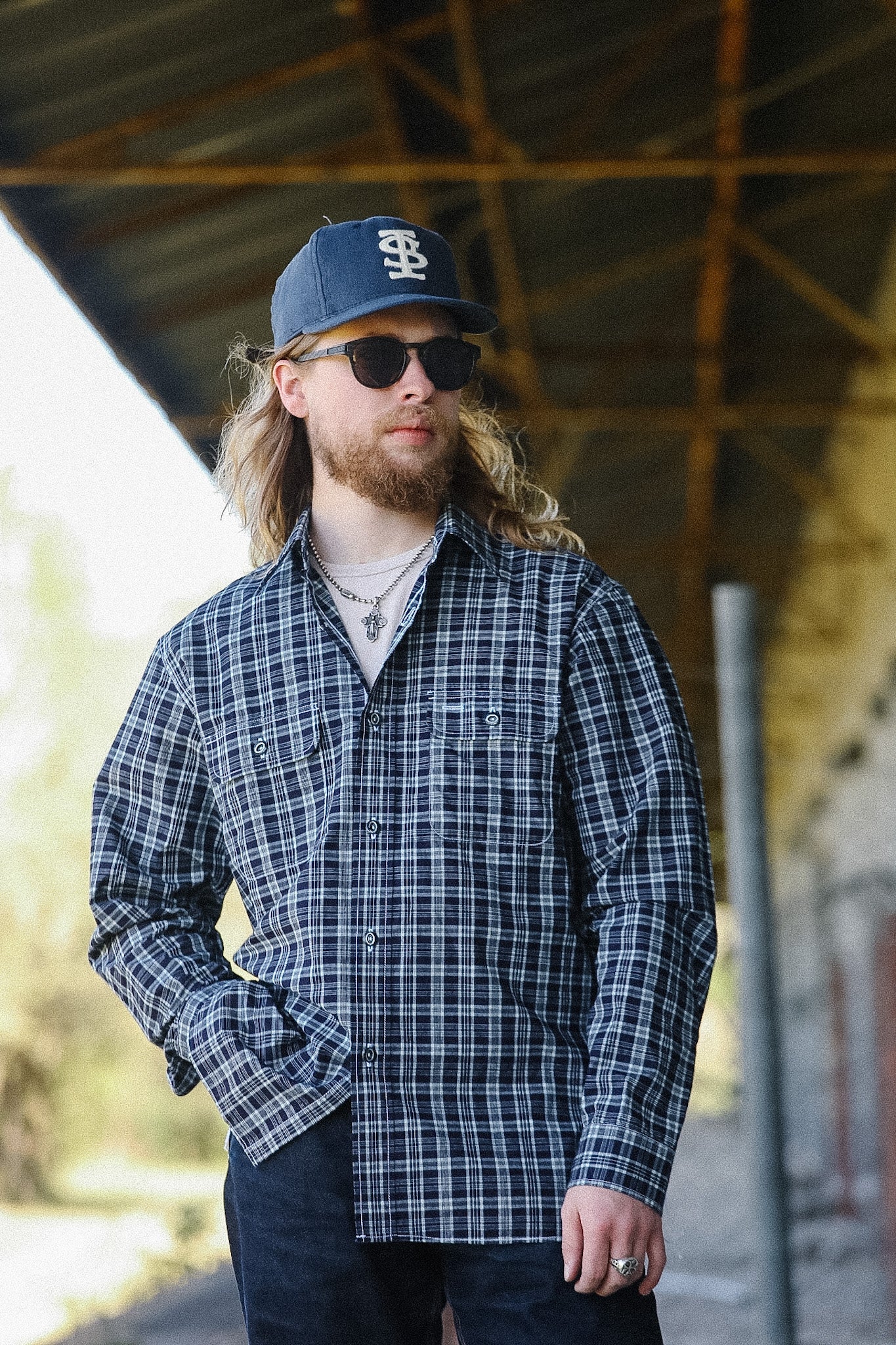 Man wearing a blue plaid shirt and cap with sunglasses, standing outdoors.
