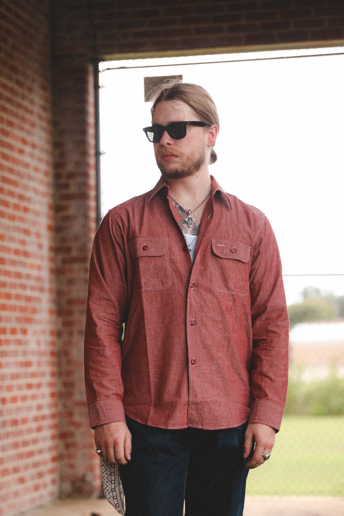 A man wearing a rust-red long-sleeve button-up shirt, dark sunglasses, and a silver necklace, standing in front of a brick wall and a bright window.