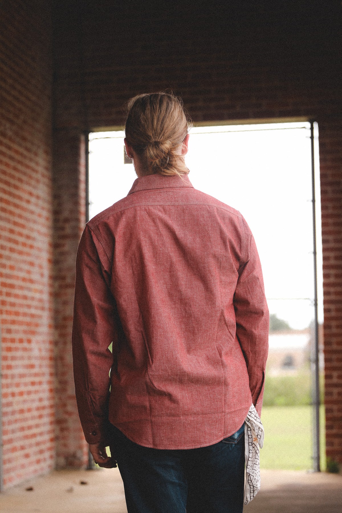 A man viewed from behind wearing a rust-red long-sleeve button-down shirt and dark jeans, standing in a brick corridor looking out through a doorway.