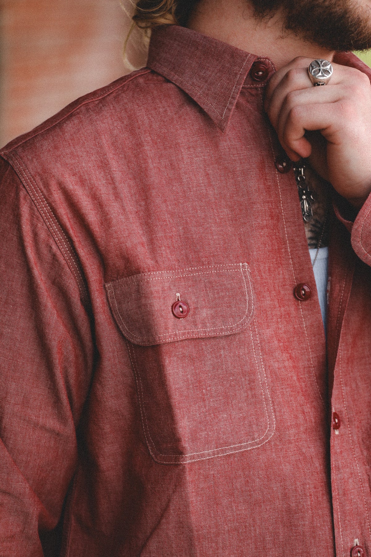 Close-up of a person wearing a rust-red or terracotta button-up shirt with a chest pocket and visible contrast stitching. A hand with a silver ring is near the collar area.