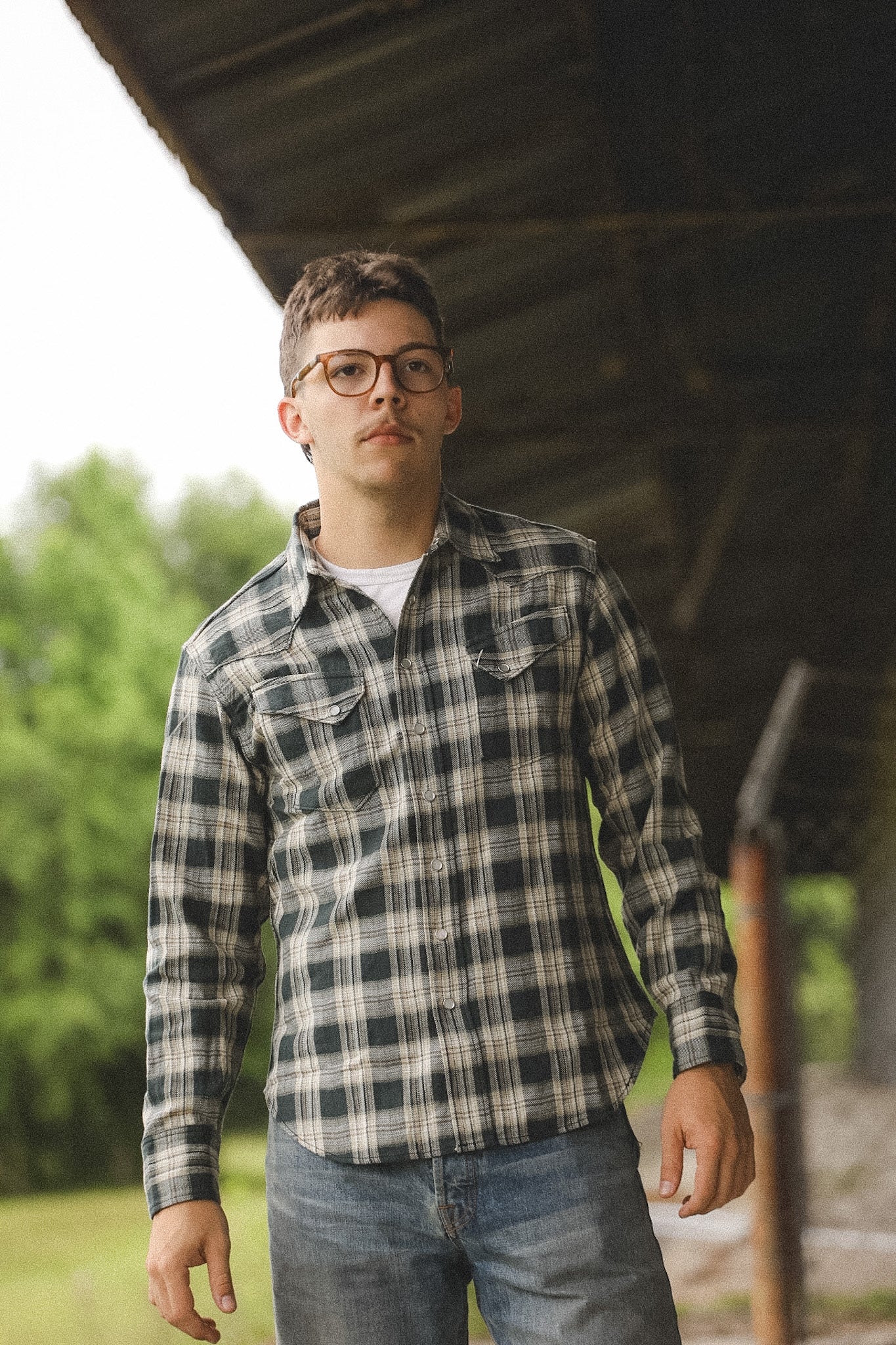 Man wearing a plaid shirt and glasses standing outdoors with trees in the background