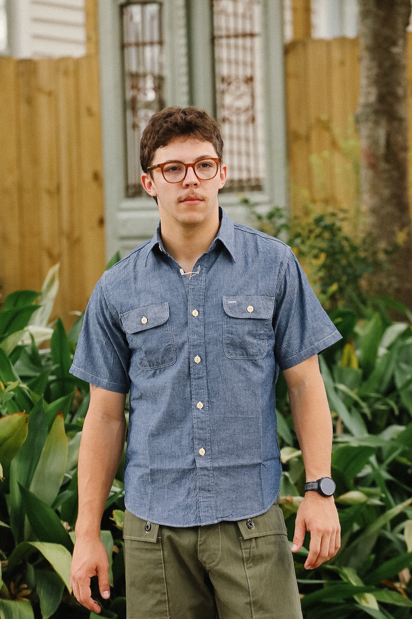 Person wearing a blue shirt and green pants standing outdoors with plants and a wooden fence in the background