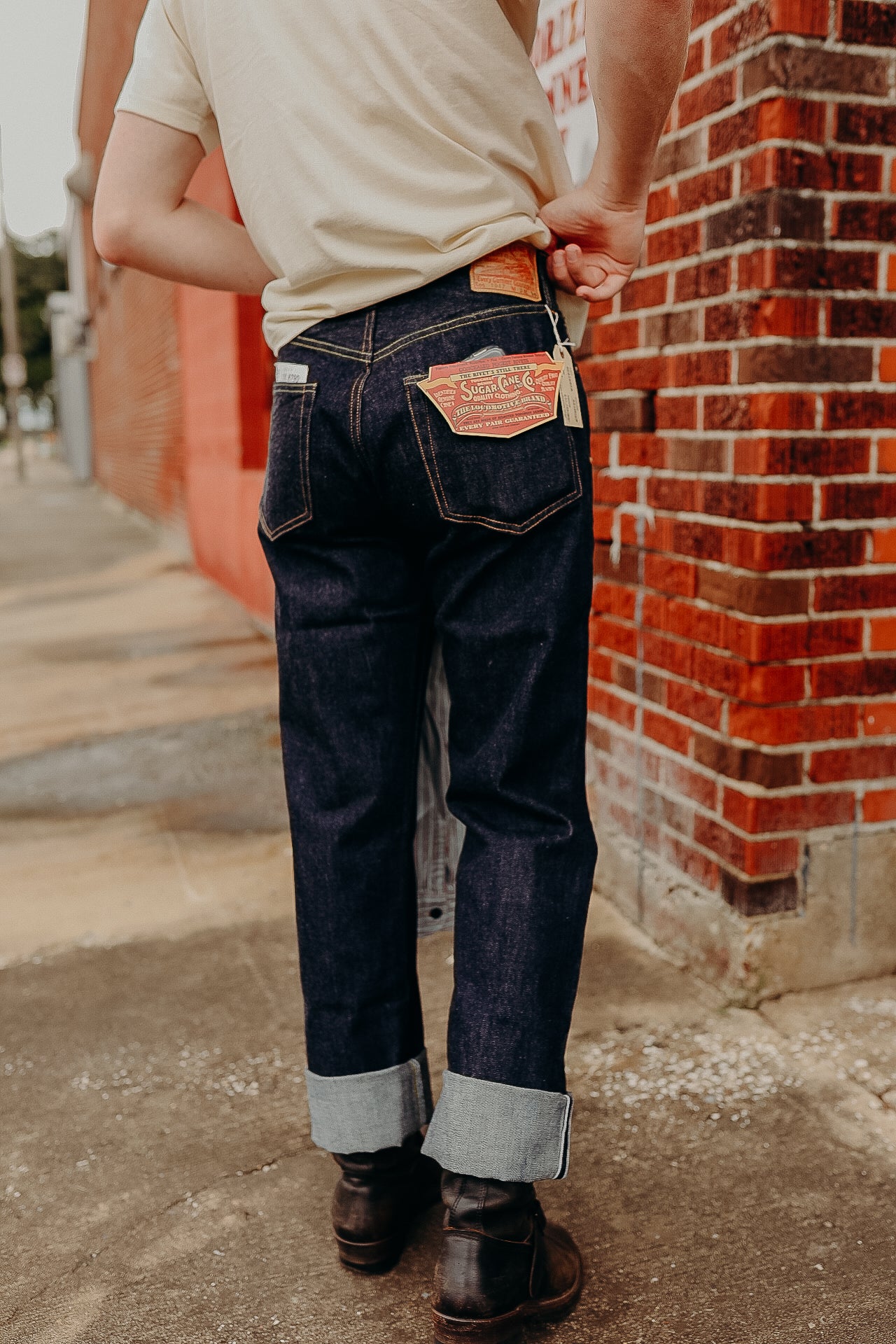 Person wearing dark raw denim jeans with cuffs and dark leather boots standing by a red brick wall