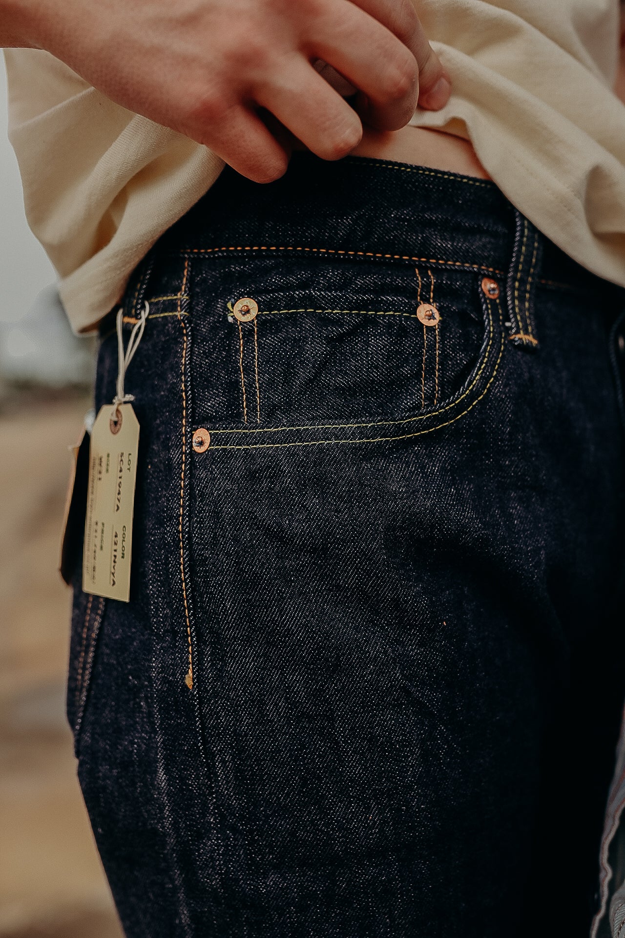 Close-up of person wearing dark raw denim jeans with tags attached, showcasing detailed stitching and copper rivets