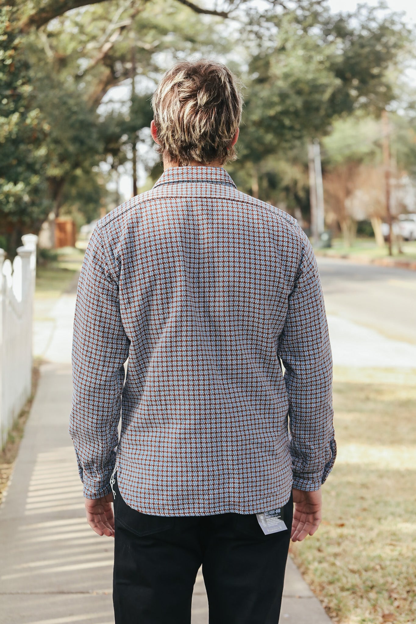 Man wearing a plaid shirt walking on a sidewalk with trees and grass in the background