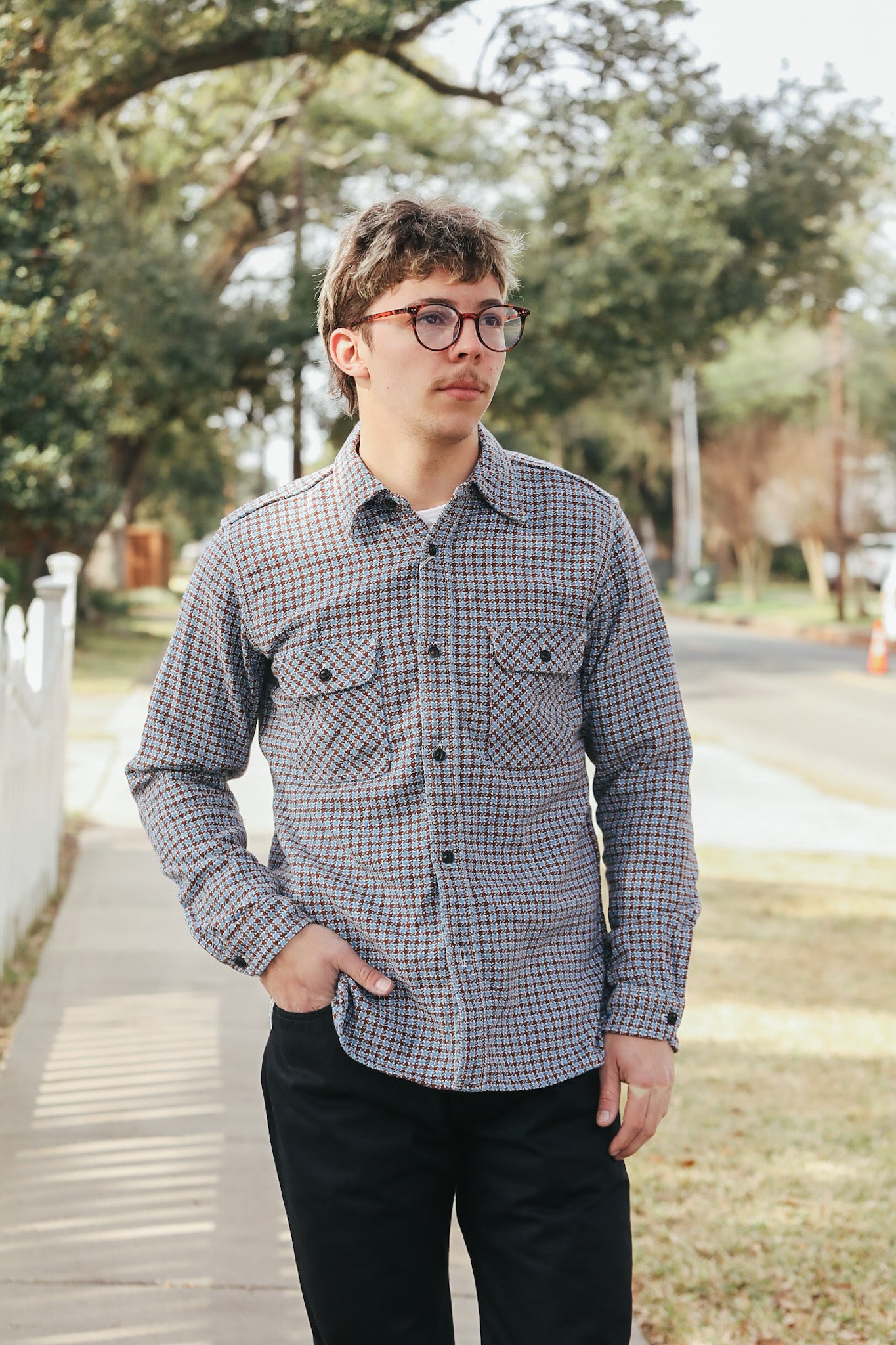 Man wearing a checkered shirt and glasses standing outdoors with trees in the background