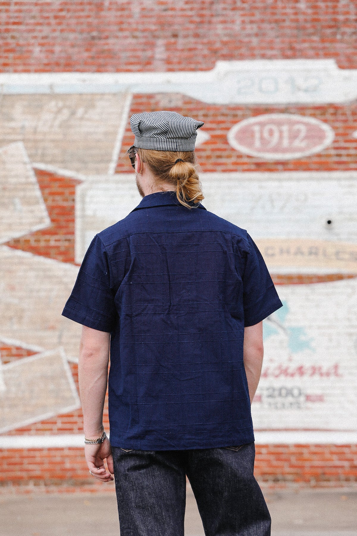 A man viewed from behind wearing a navy blue short-sleeve button-up shirt, dark jeans, and a gray textured flat cap, standing in front of a brick wall with a painted mural.