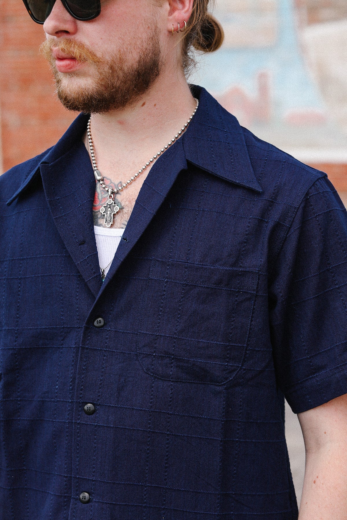 A close-up of a man wearing a dark navy blue short-sleeve button-up shirt with a textured grid pattern, layered over a white undershirt and accessorized with a silver necklace.