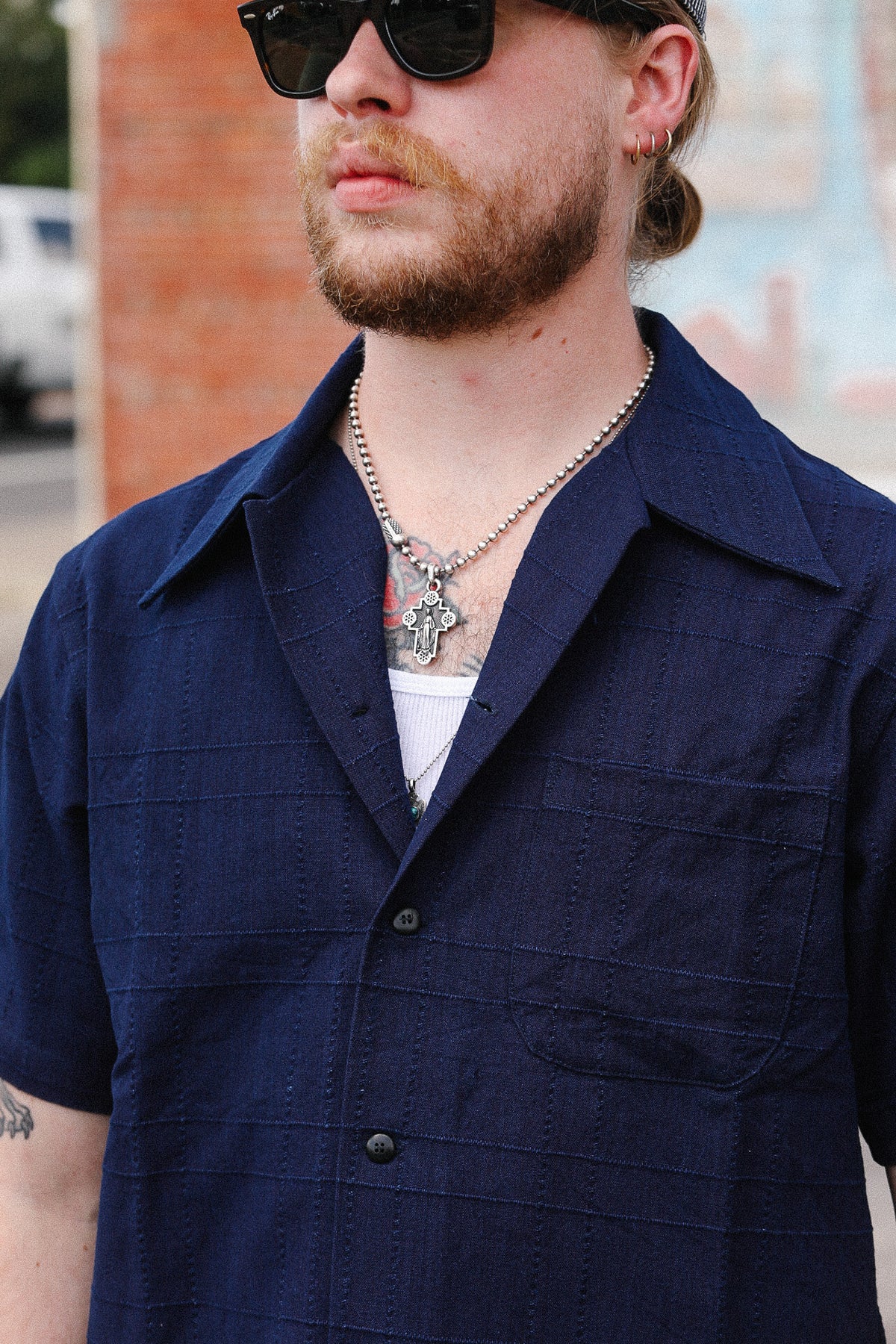A man wearing a dark navy blue short-sleeve button-up shirt with a subtle textured grid pattern, styled with a white undershirt, silver chain necklace, and black sunglasses.