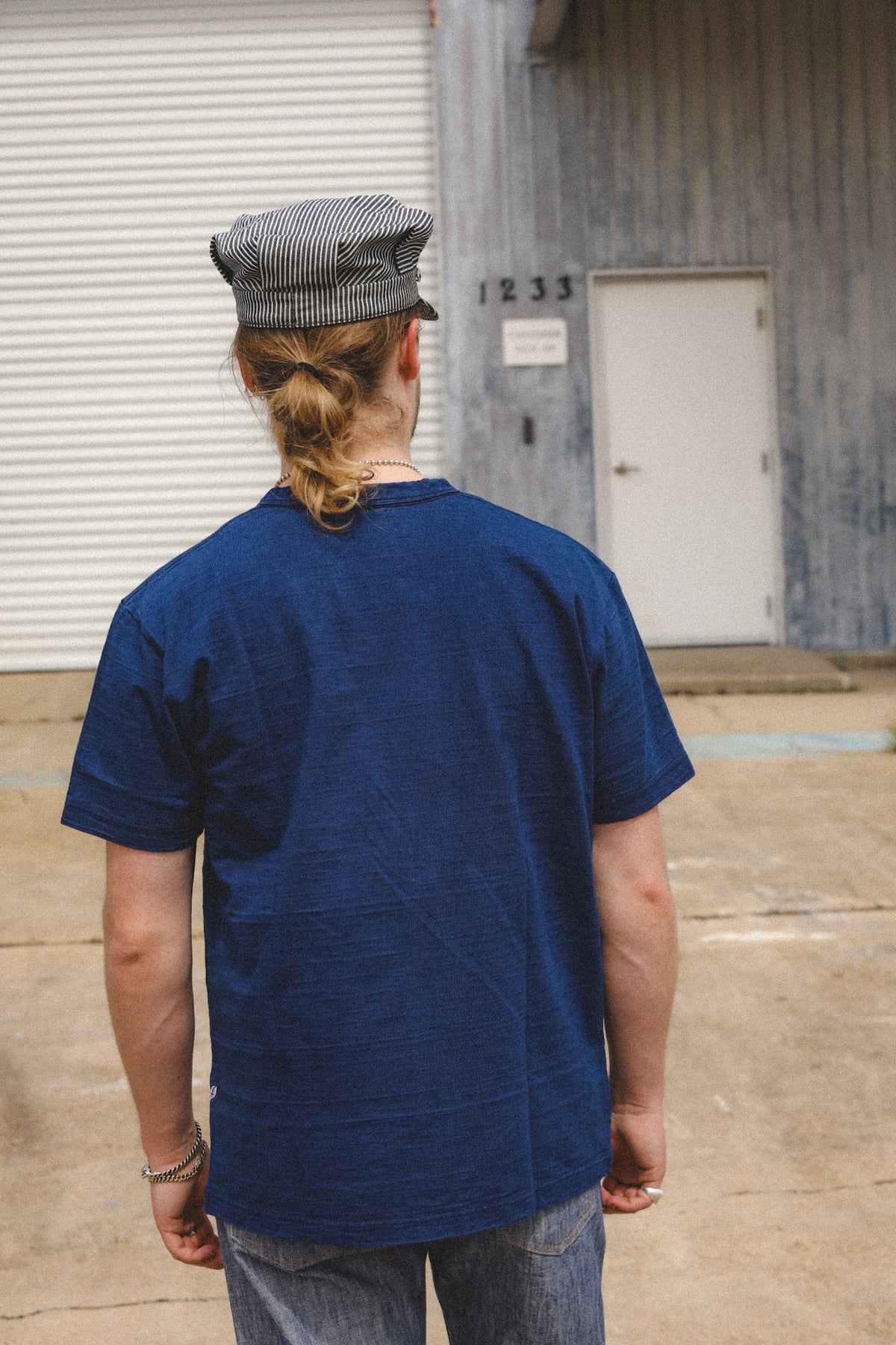 A person viewed from behind wearing a dark blue short-sleeve t-shirt, a gray and white striped flat cap, and light blue jeans, standing outdoors in front of a gray building with a white door.