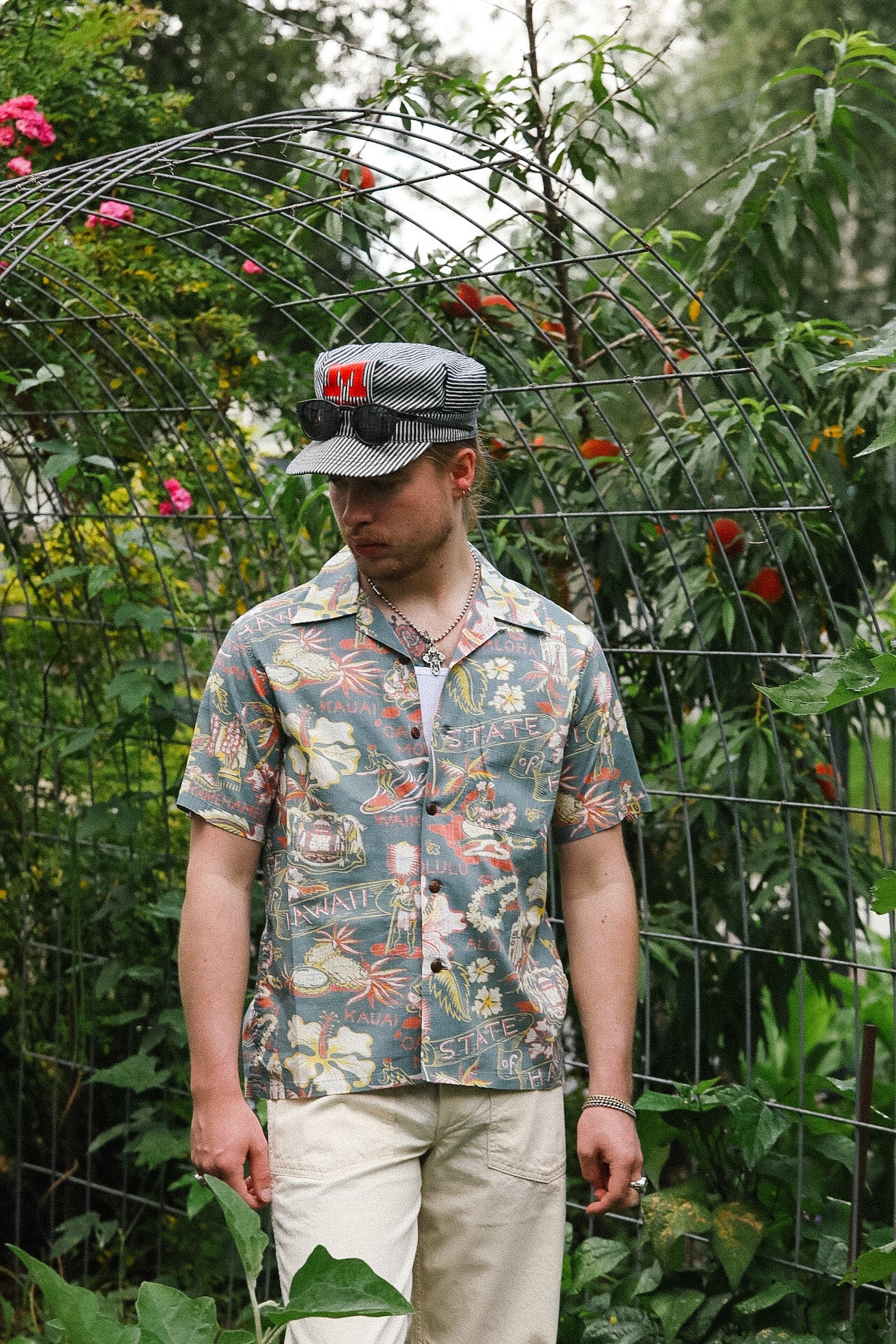Man wearing a patterned shirt and cap standing in front of a wire fence with plants and flowers.
