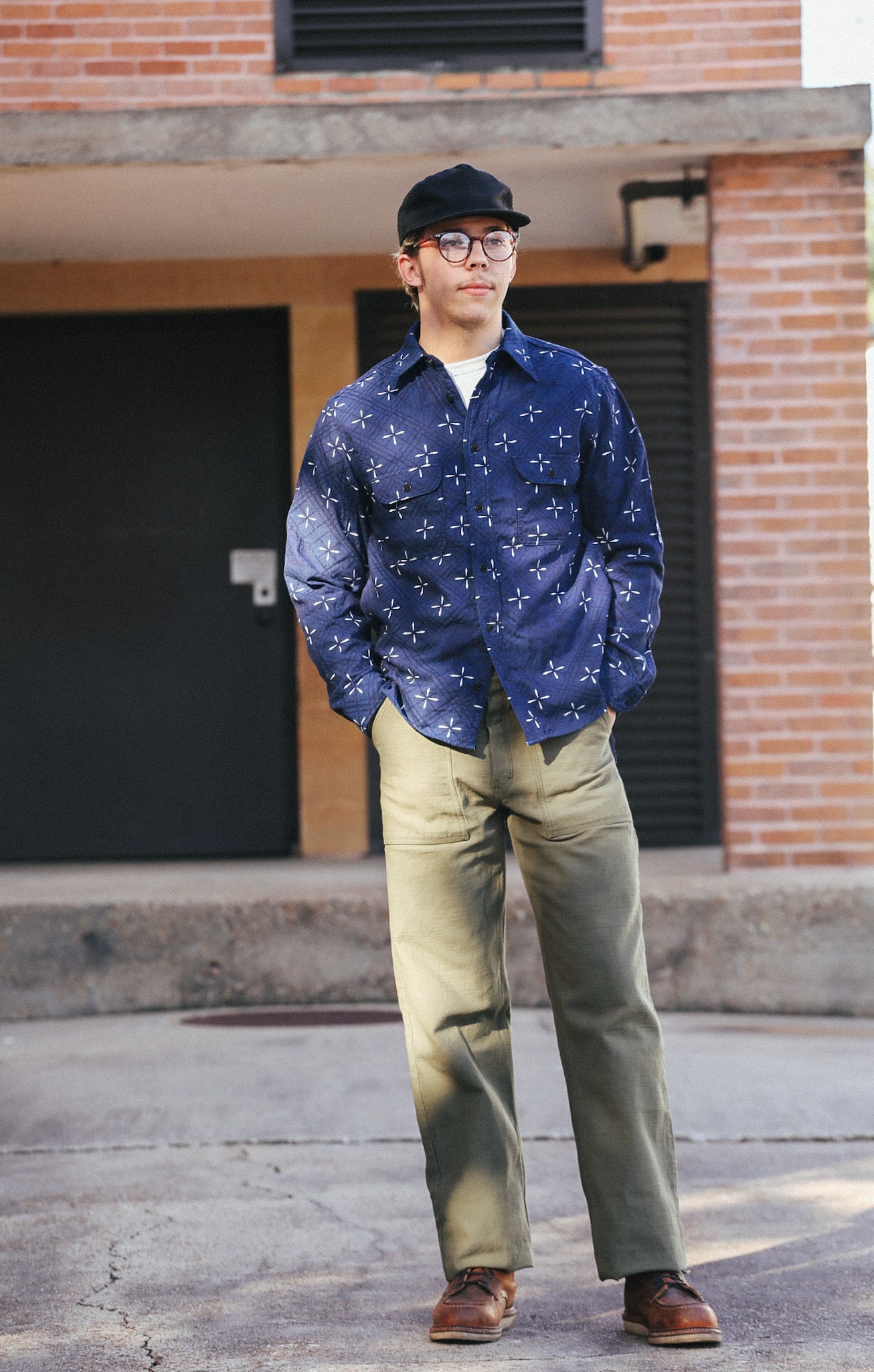 Man wearing indigo jacquard work shirt with goldfish pattern, beige pants, brown shoes, and black cap in urban setting