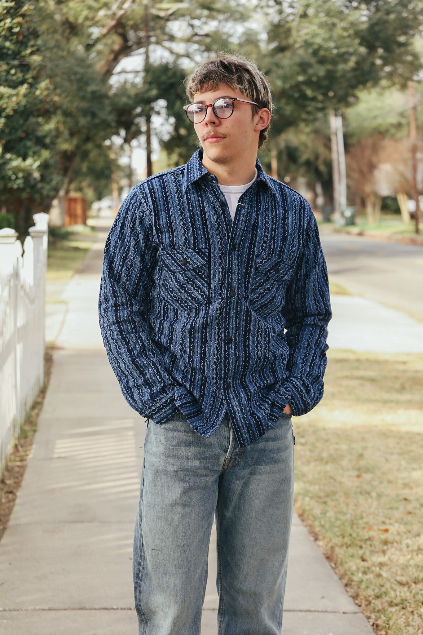 Person wearing a blue patterned shirt and gray jeans standing on a sidewalk with trees and grass in the background.