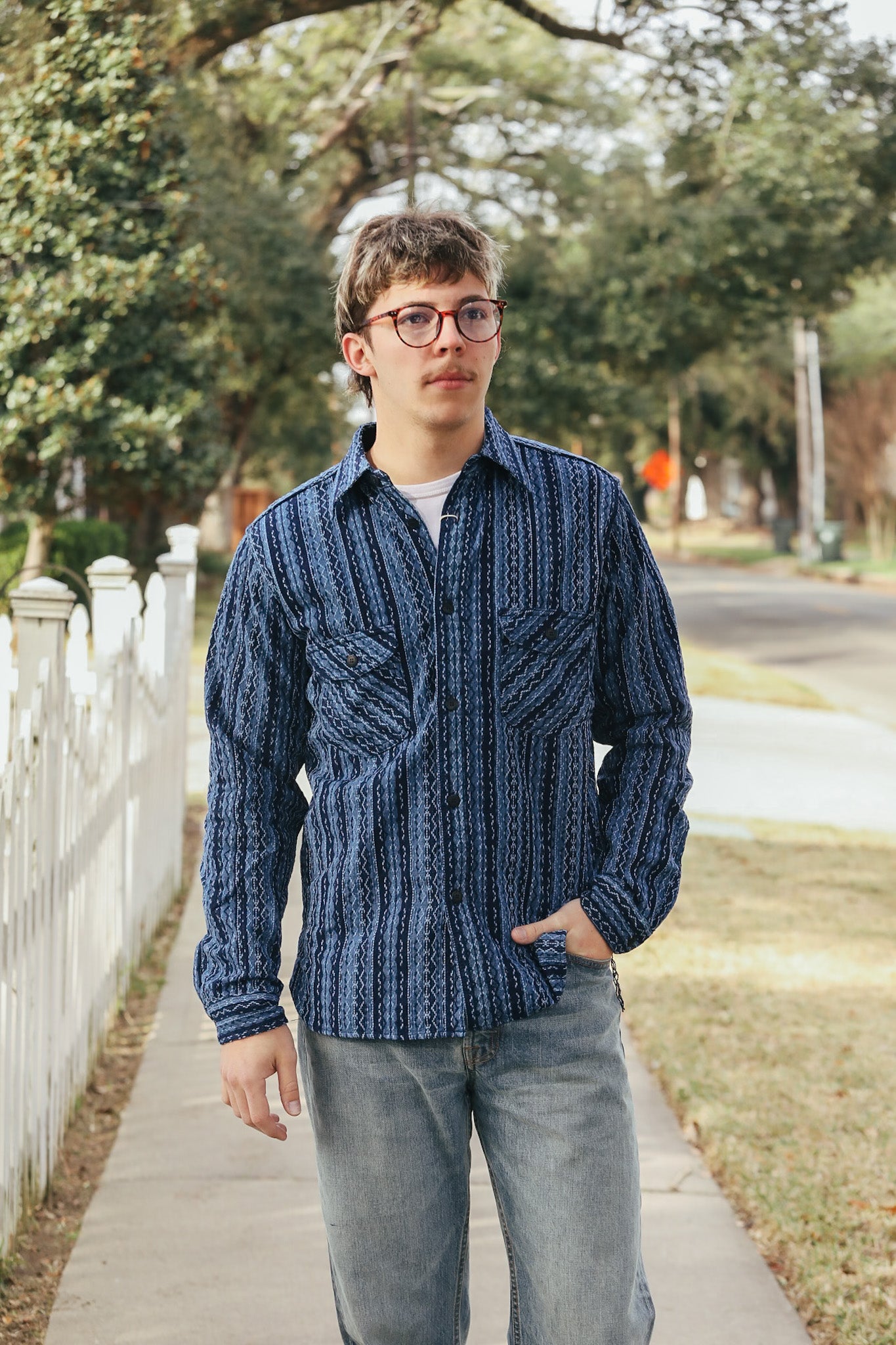 Man wearing a blue patterned shirt and glasses standing on a sidewalk with trees and a white fence in the background.