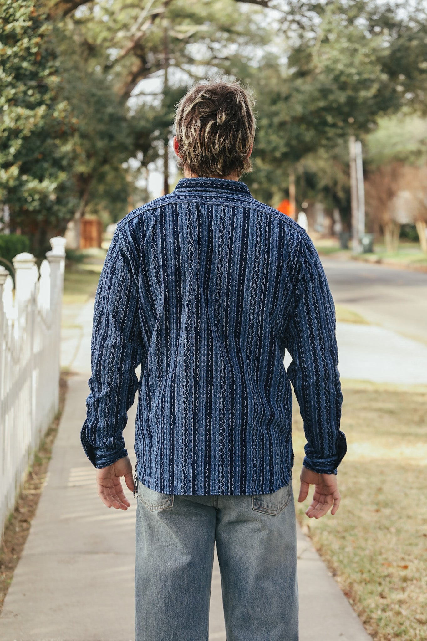 Man wearing a blue patterned shirt walking away on a sidewalk with trees and a white fence in the background.