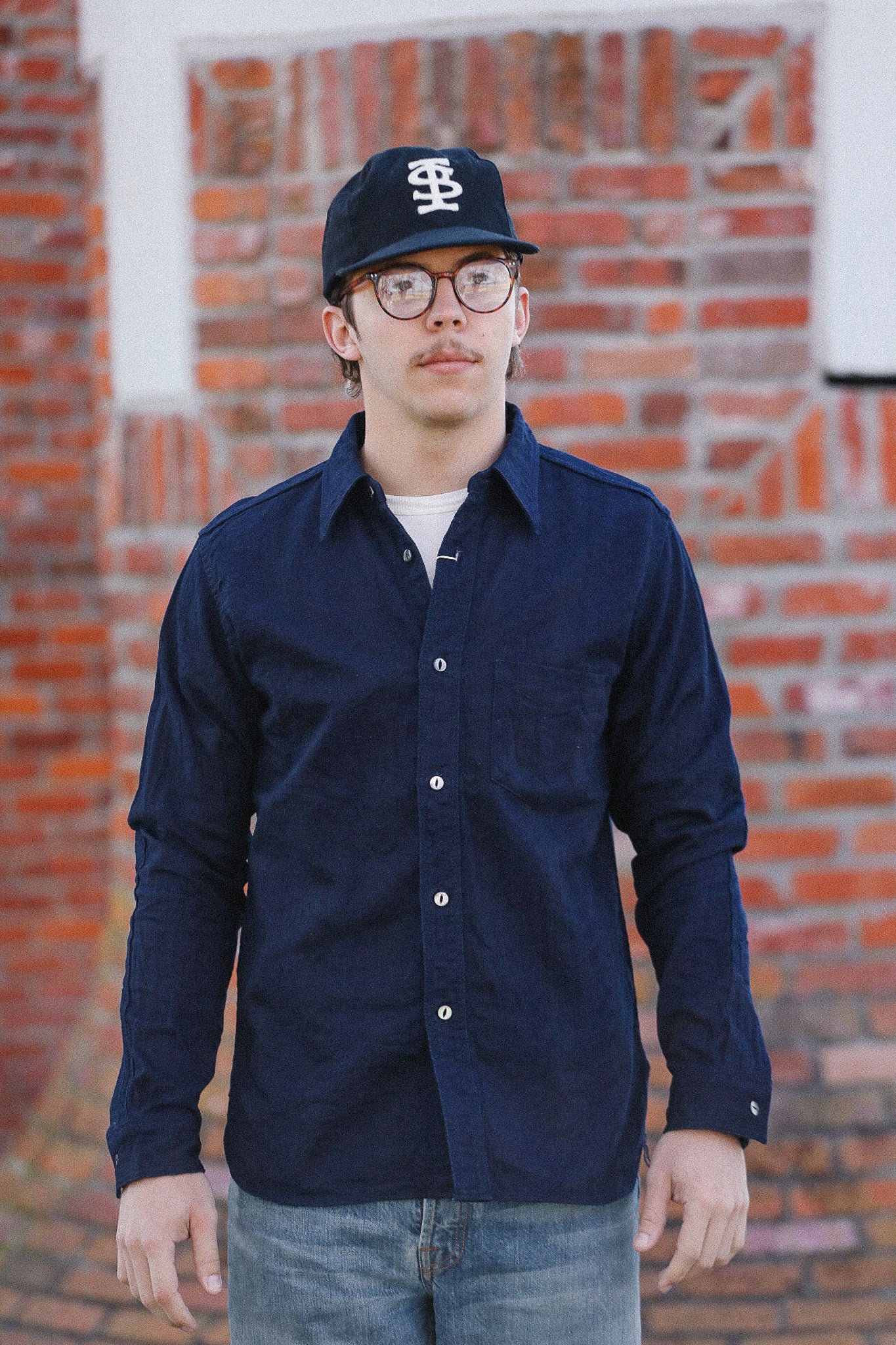 Person wearing a navy button-up shirt and cap in front of a brick wall