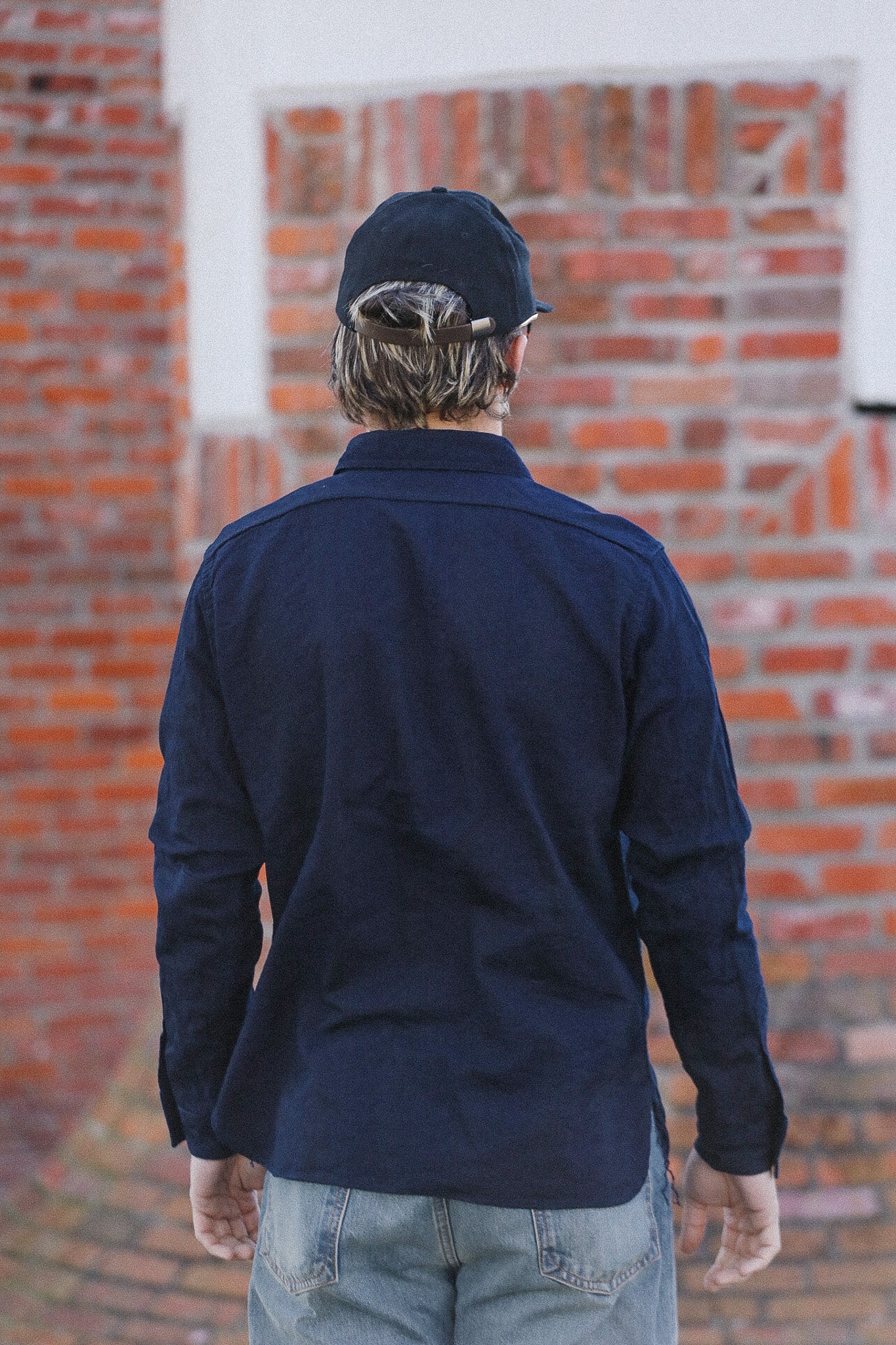Person wearing a navy blue shirt and cap standing against a brick wall.