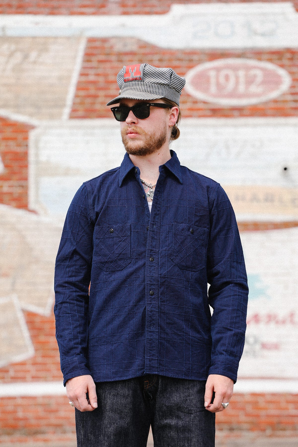 A man wearing a dark navy blue long-sleeve button-up shirt, dark jeans, a gray cap, and sunglasses, standing in front of a brick wall with painted lettering.