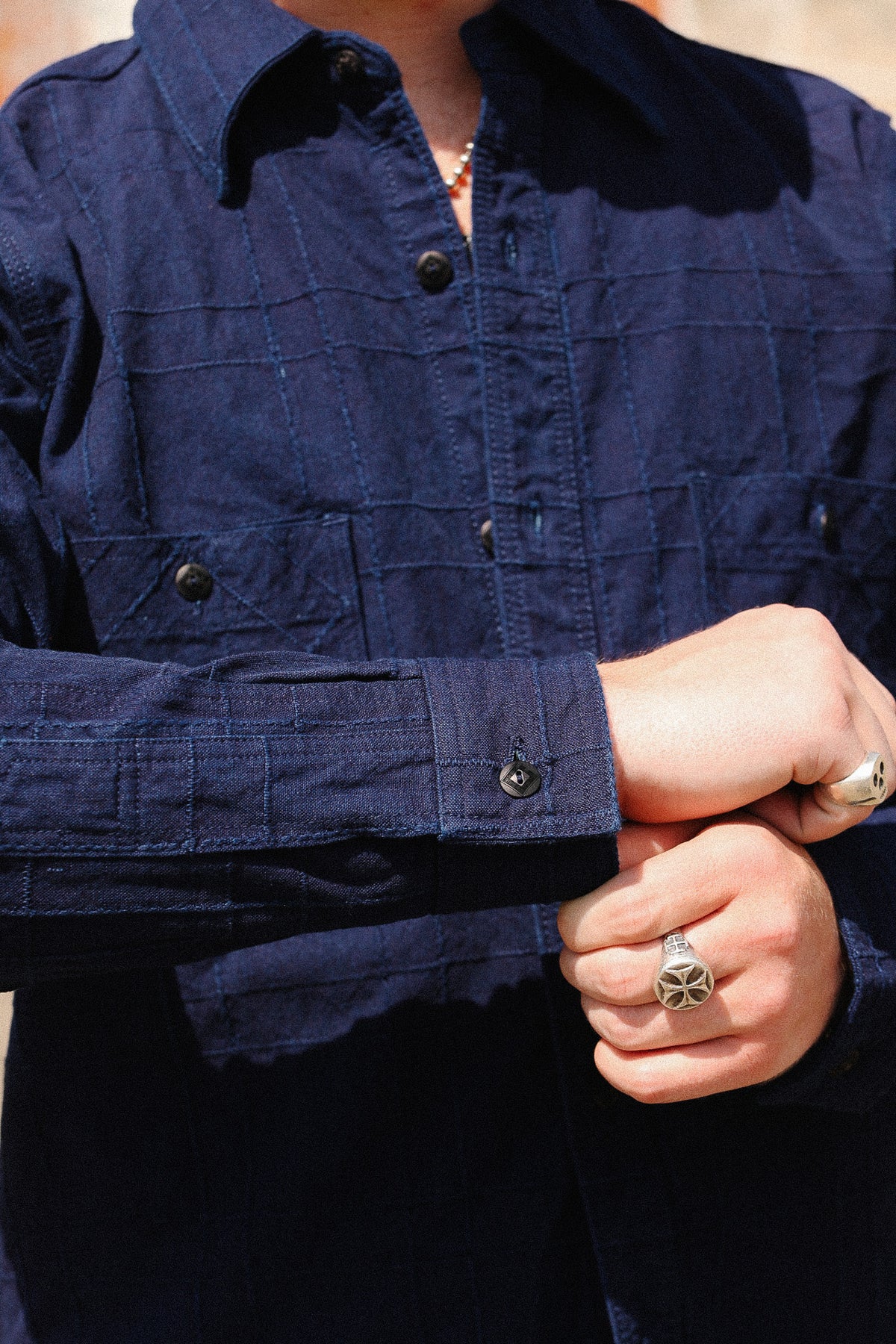 Close-up of a person wearing a dark navy long-sleeve button-up shirt with a subtle grid texture, adjusting the cuffs with silver rings visible on their fingers.