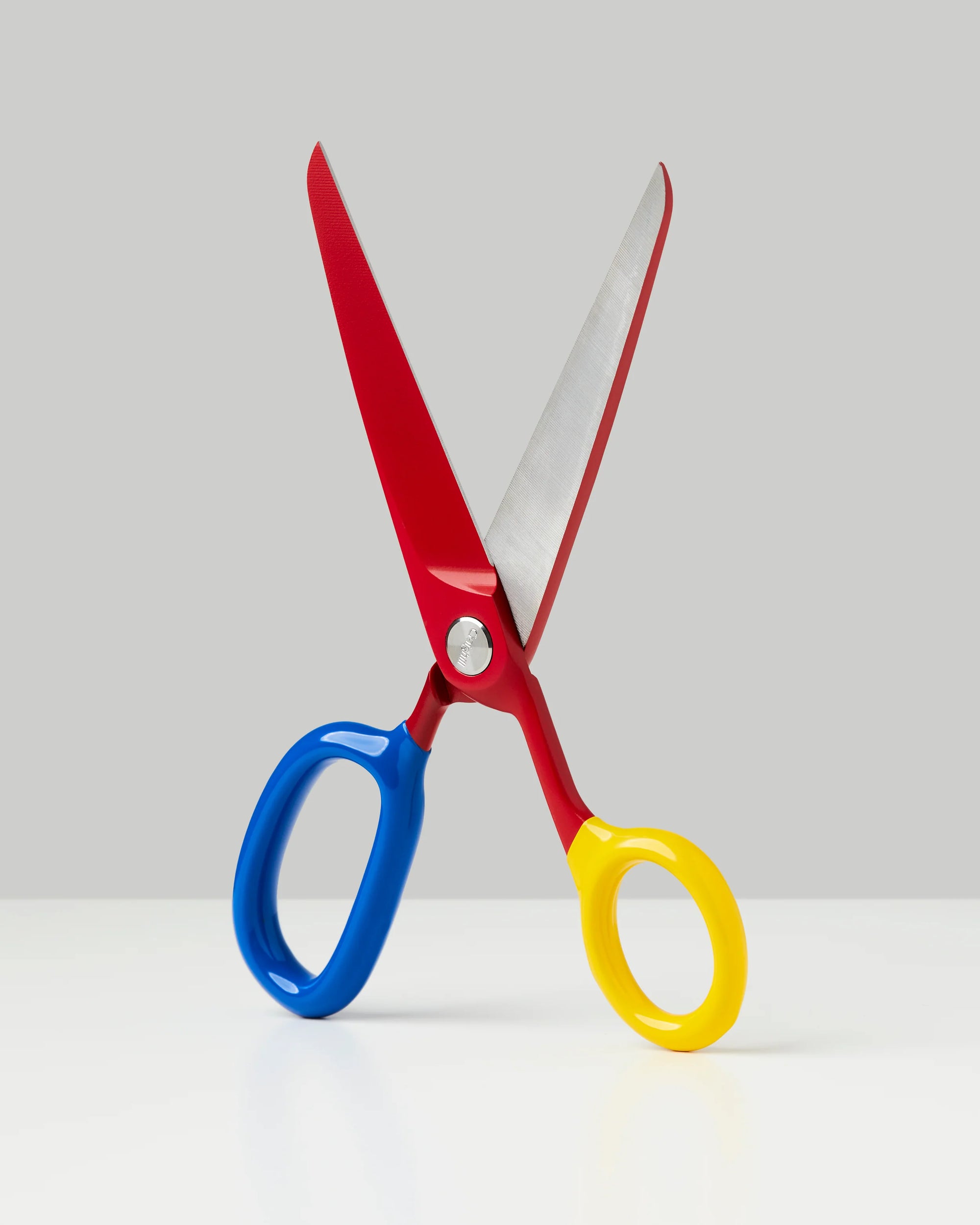 Scissors with red blades, blue and yellow ergonomic handles on white surface against gray background