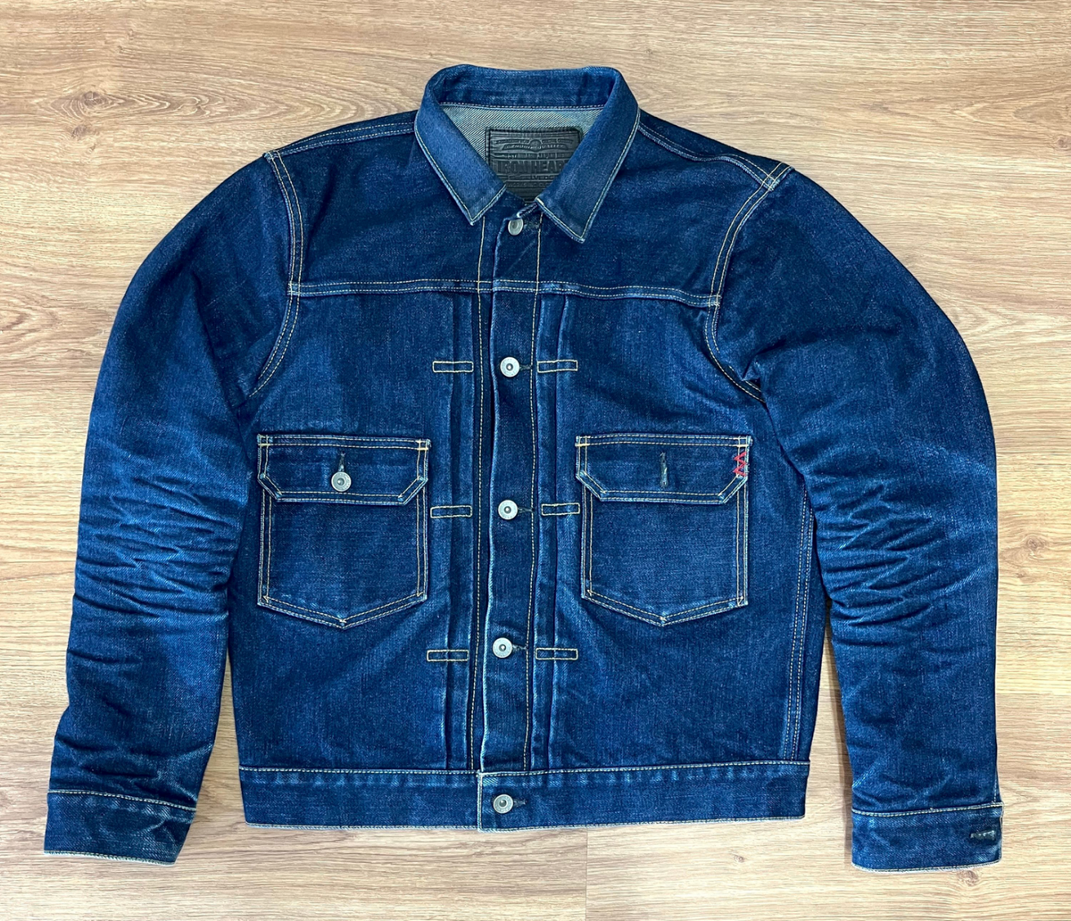 A dark blue denim trucker jacket with contrast stitching, laid flat on a light brown wooden floor. The jacket features a button-up front, two chest pockets with button closures, and a classic collar.