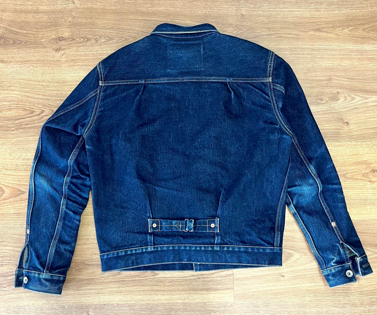 A dark blue denim jacket shown from the back, laid flat on a light brown wooden floor.