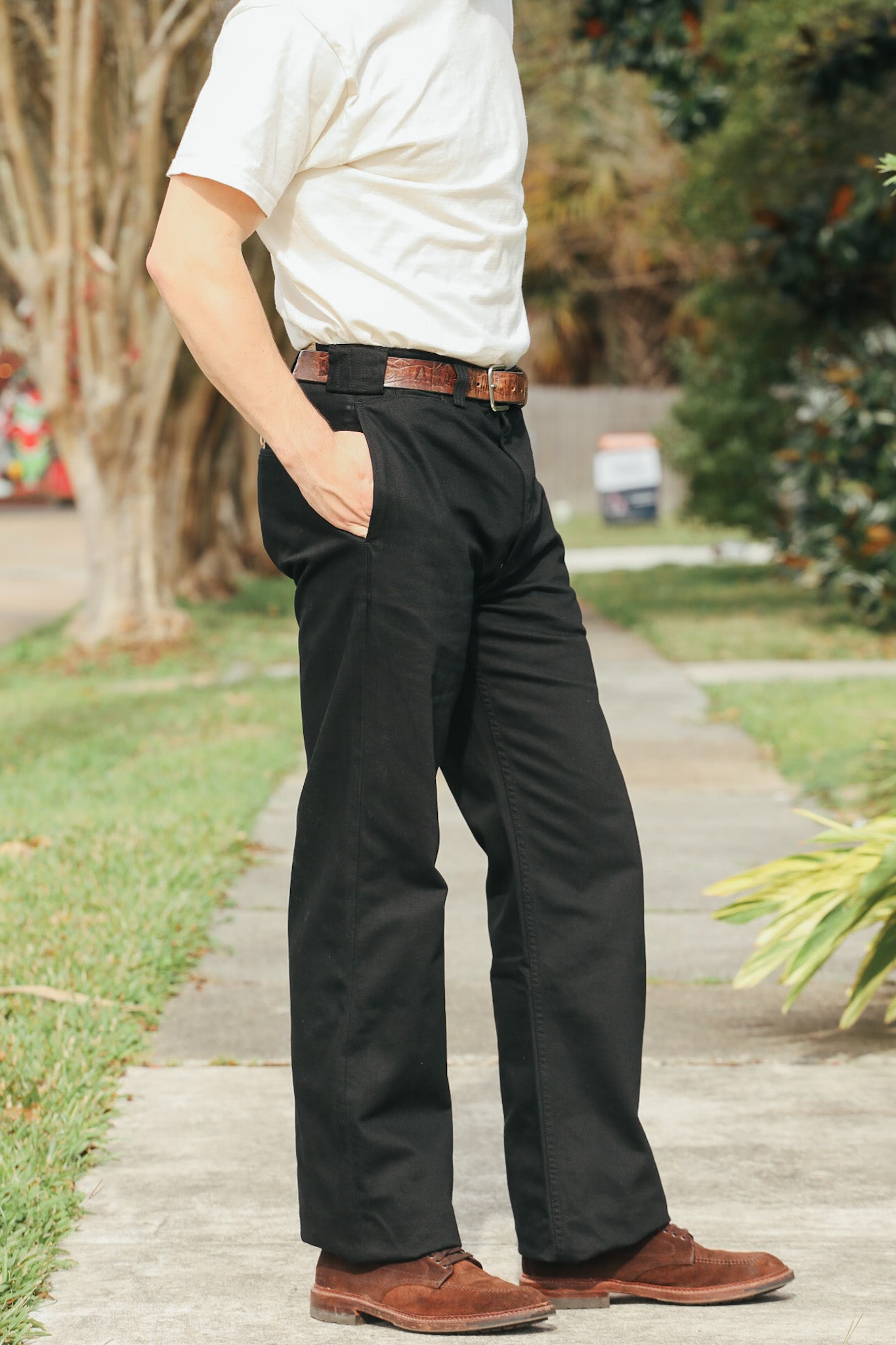 Person wearing a white shirt, black pants, and brown shoes on a sidewalk with greenery in the background.