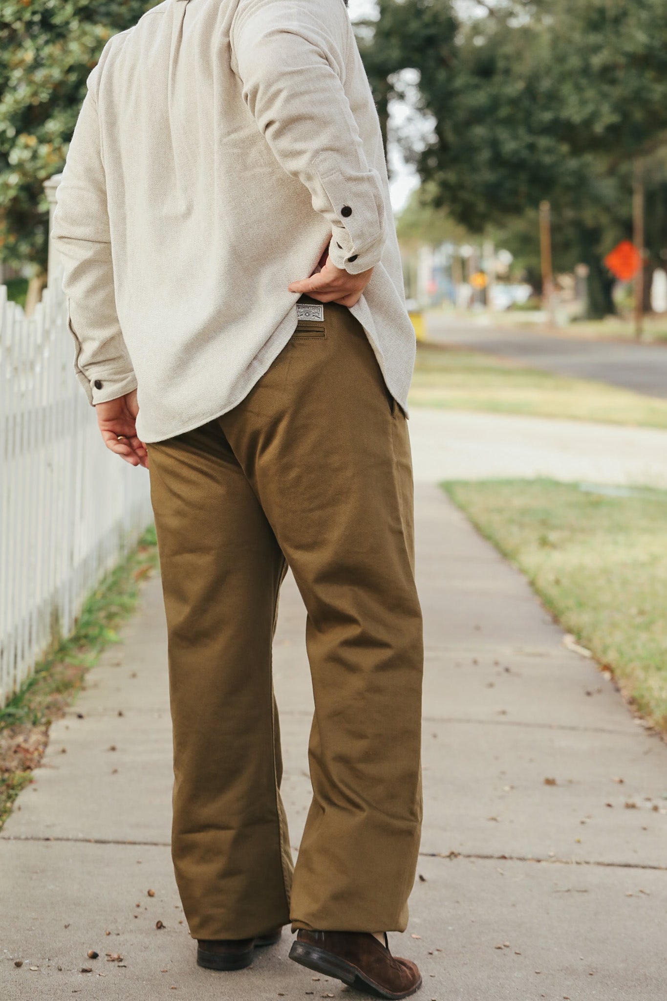 Person wearing a beige jacket and brown pants on a sidewalk with trees and a stop sign in the background.