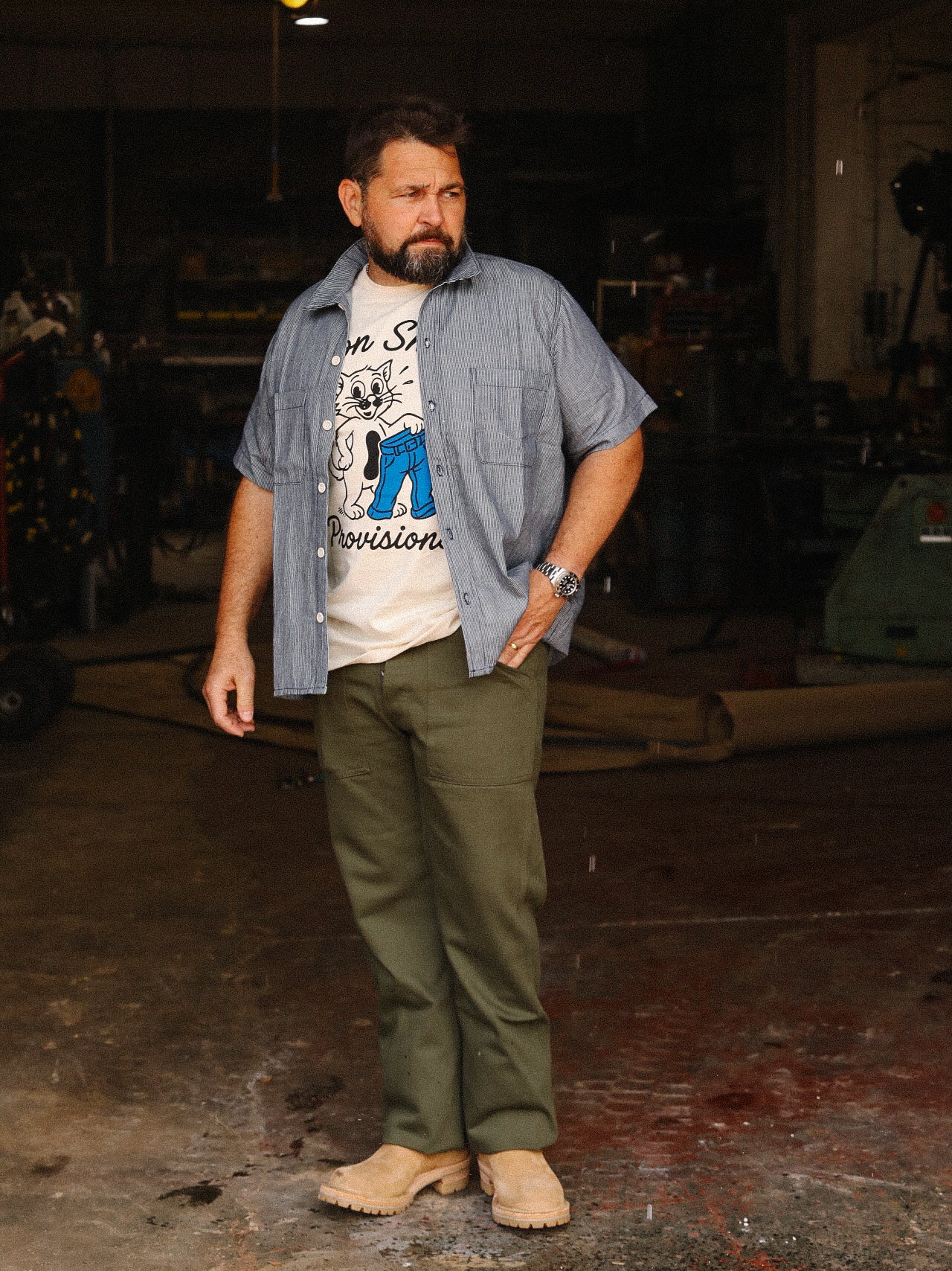 Man wearing short sleeve rainbow nep hickory stripe shop shirt, graphic tee, olive pants, and beige boots in workshop