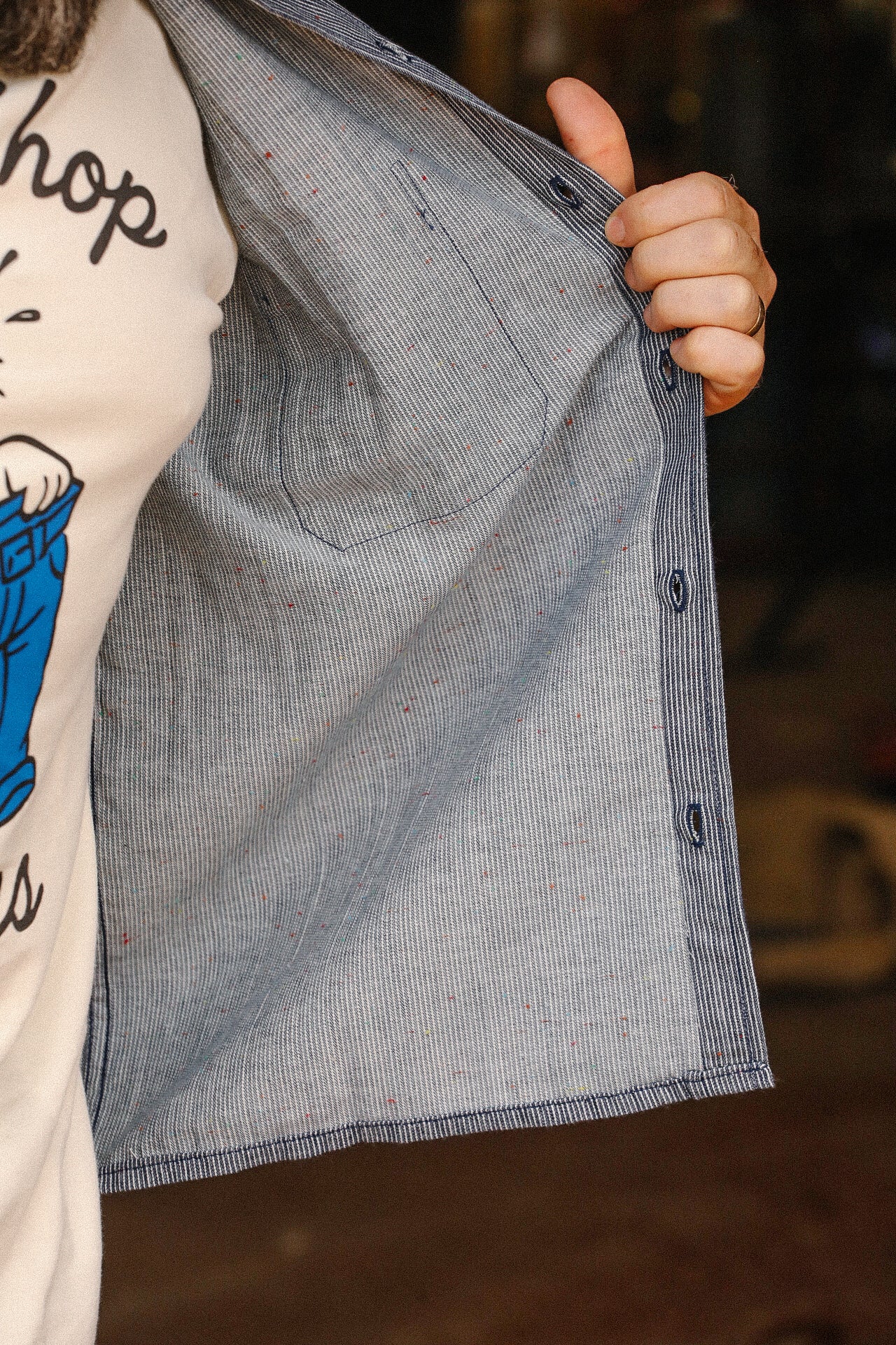 Close-up of a hand holding open a blue and white striped shirt with rainbow nep details inside, worn over a graphic tee at Iron Shop Provisions