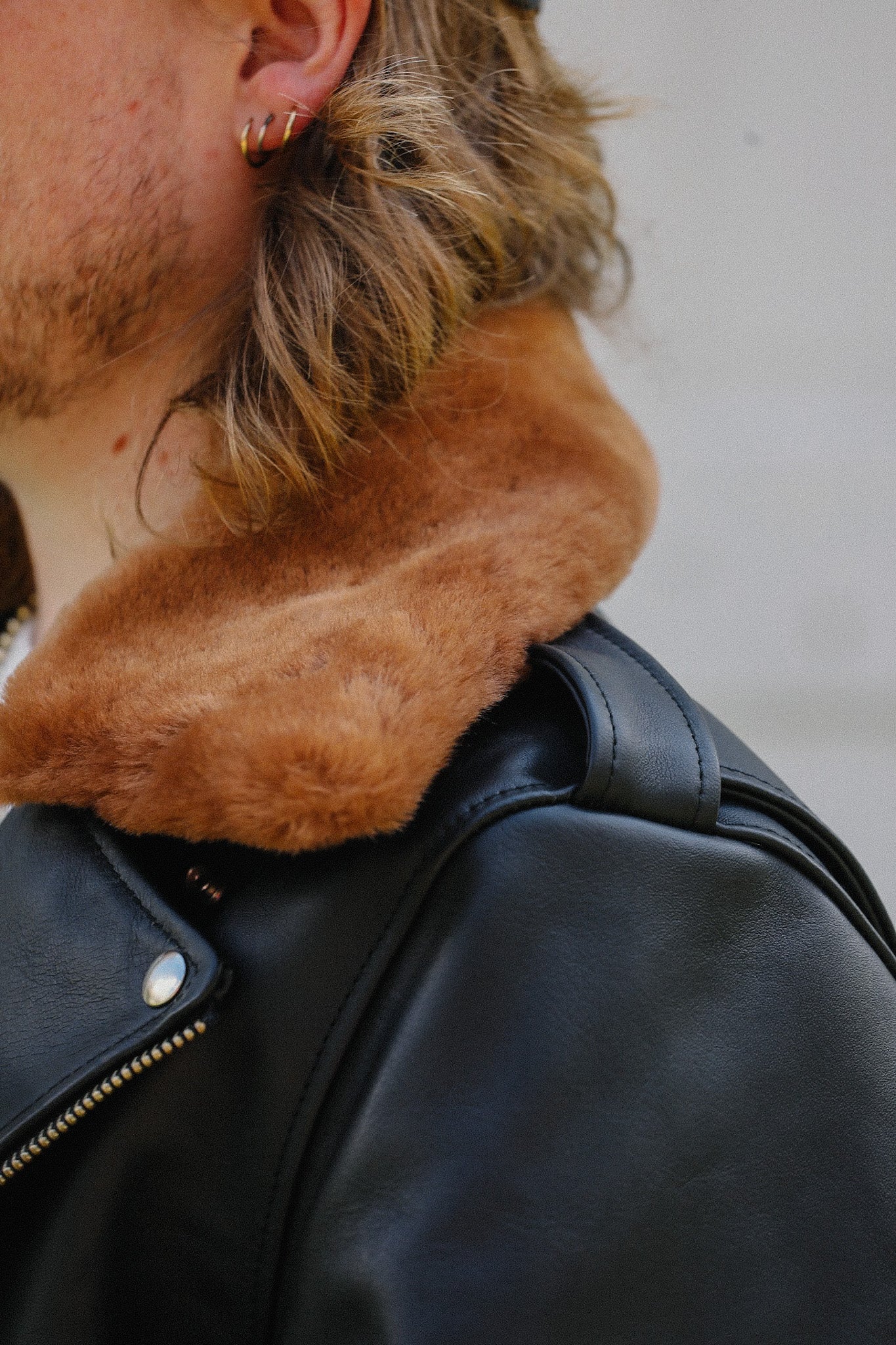 Close-up of a black leather jacket with a brown fur collar worn by a person with light hair and gold hoop earrings