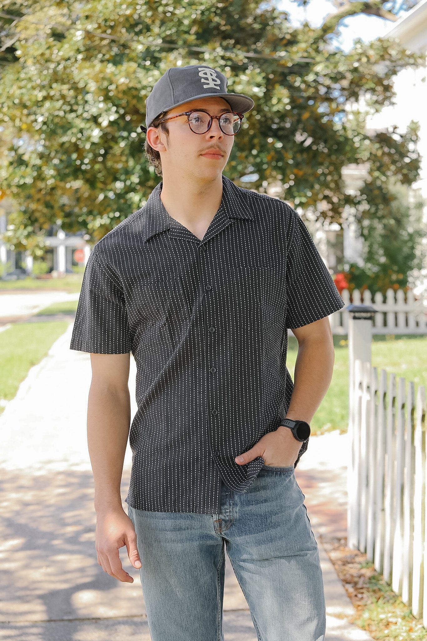 Man wearing a checkered shirt, cap, and glasses standing outdoors with trees and a white fence in the background.