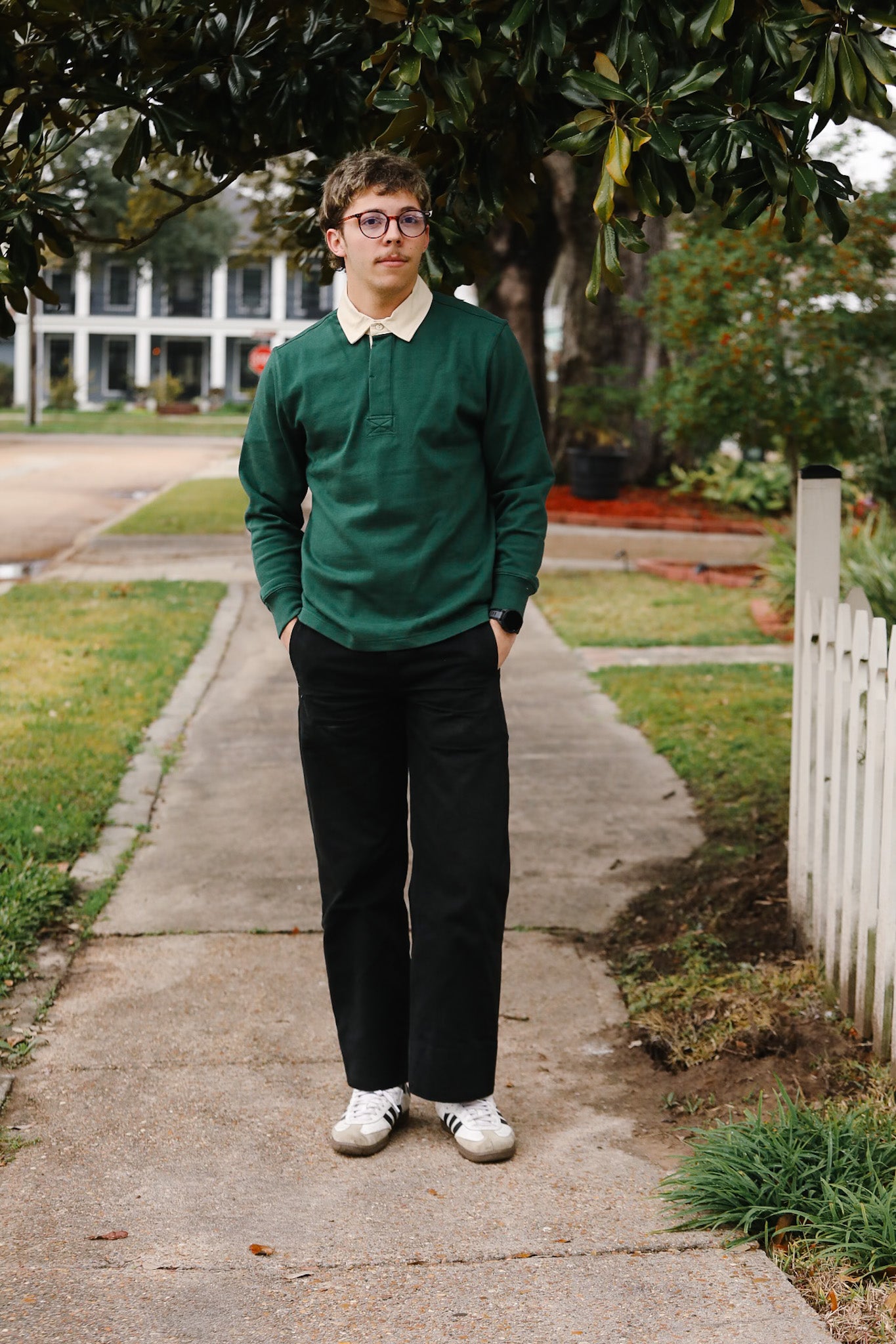 Man in a green sweater and black pants standing on a sidewalk with trees and a house in the background.