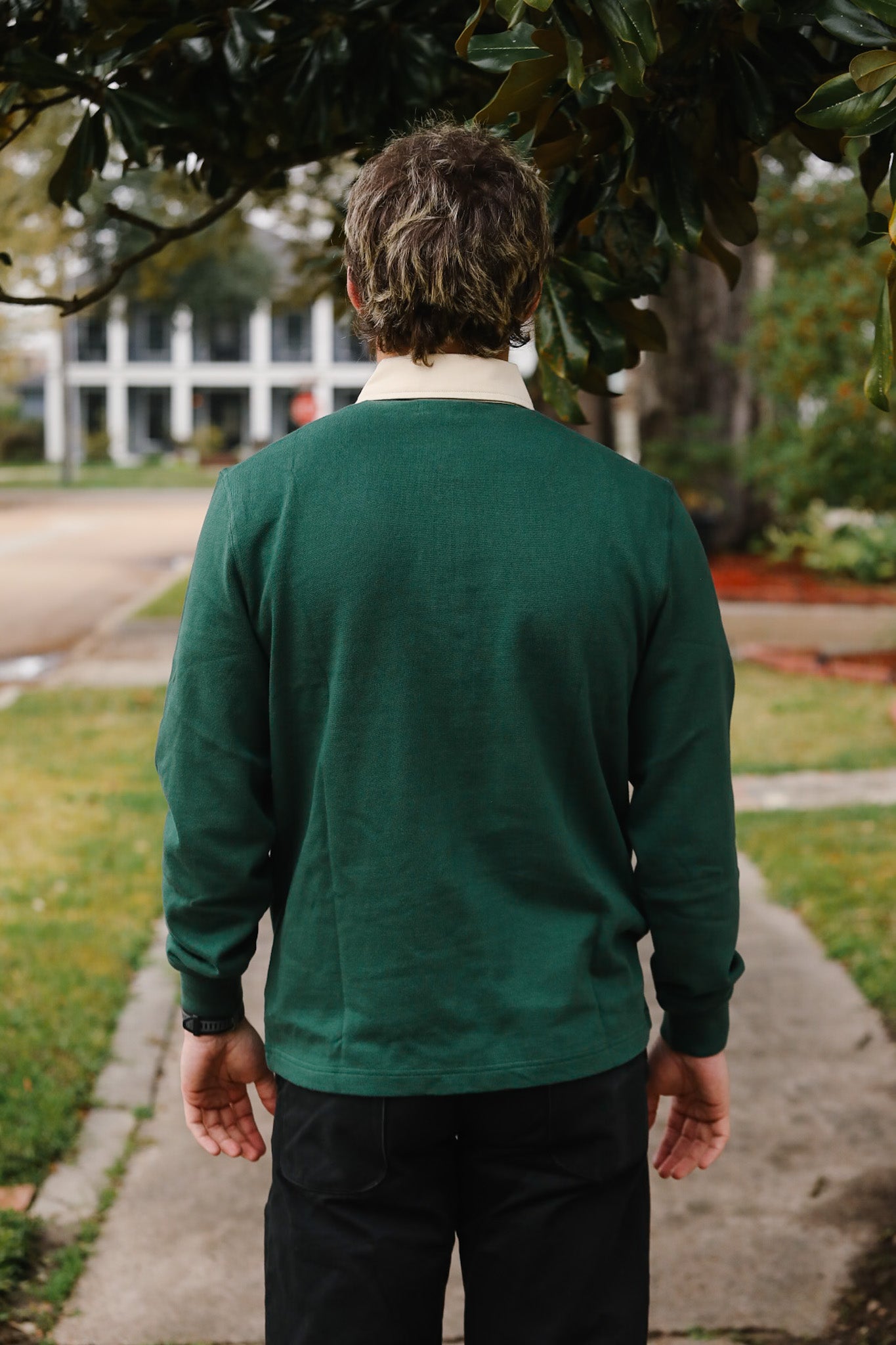 Person wearing a green jacket walking on a sidewalk with trees and buildings in the background