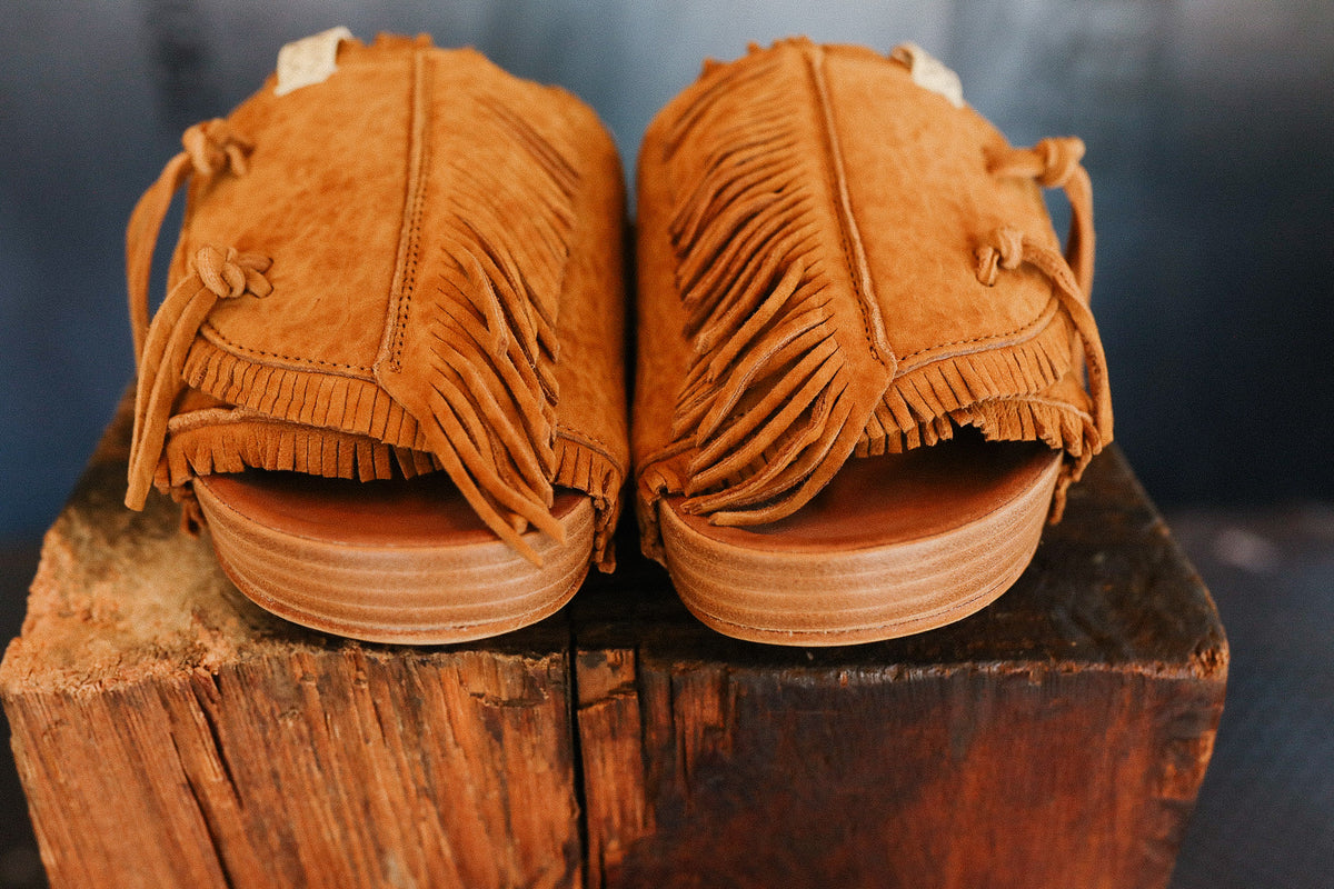 Pair of brown suede moccasins with fringes on a wooden block against a dark background