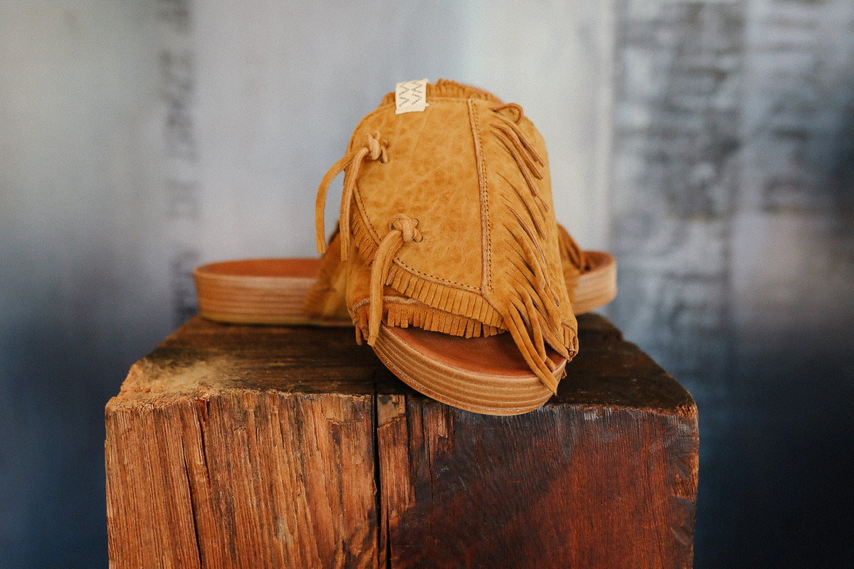 Brown suede sandal with fringes on a wooden block against a blurred background