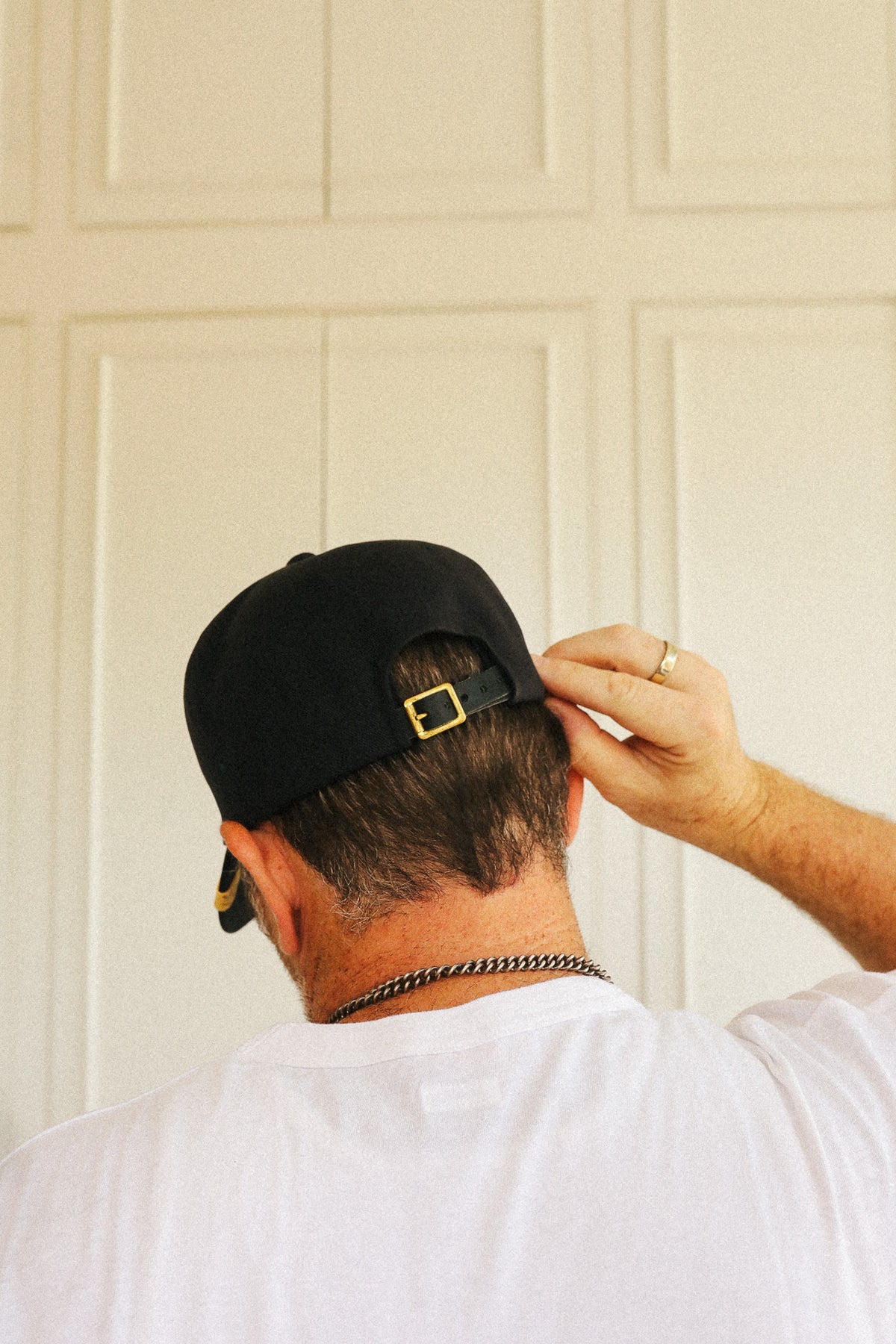 A man viewed from behind wearing a black baseball cap with a gold buckle, a white t-shirt, and a silver chain necklace, standing in front of a cream-colored paneled door.