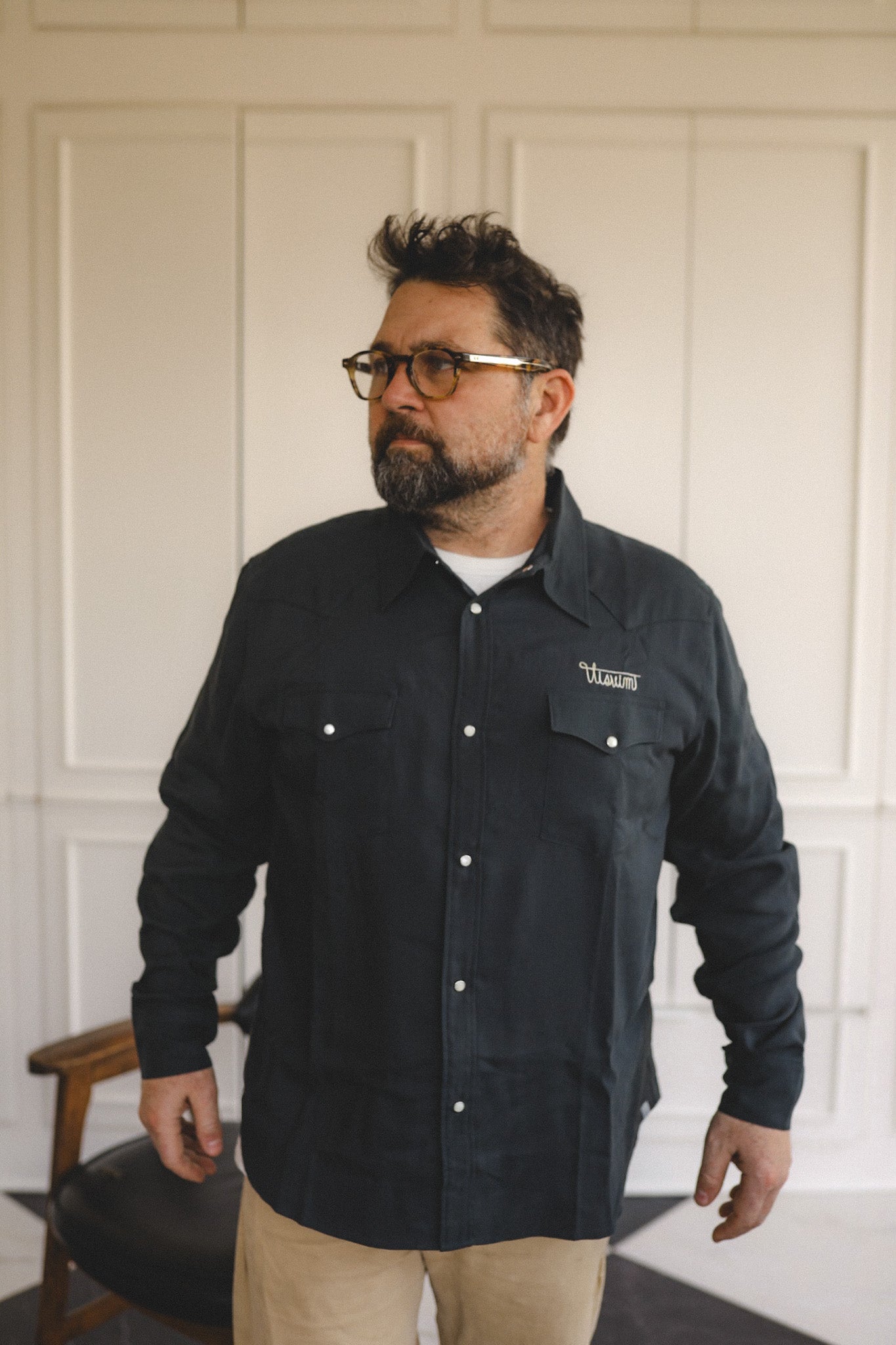 Man wearing a black jacket with a logo, standing against a white paneled wall.