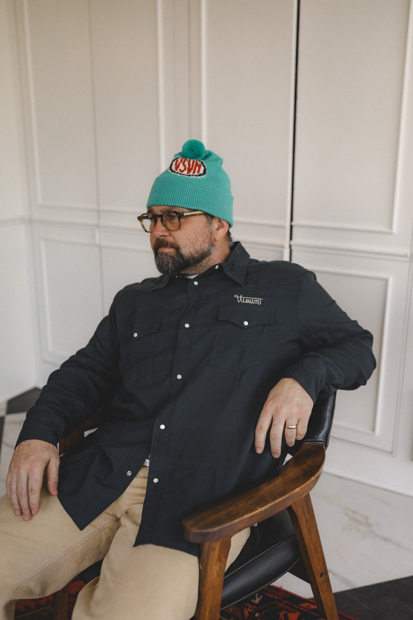Man wearing a teal beanie with a logo and a black shirt sitting in a chair.