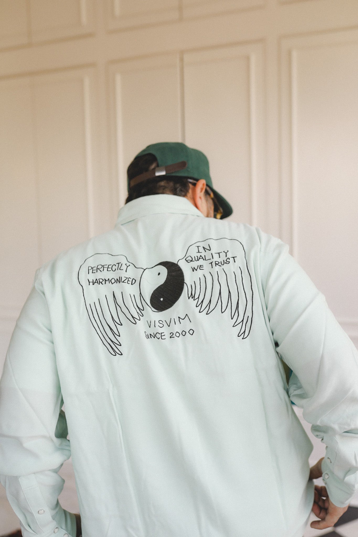 Person wearing a light blue jacket with a design and text on the back, standing against a white paneled wall.