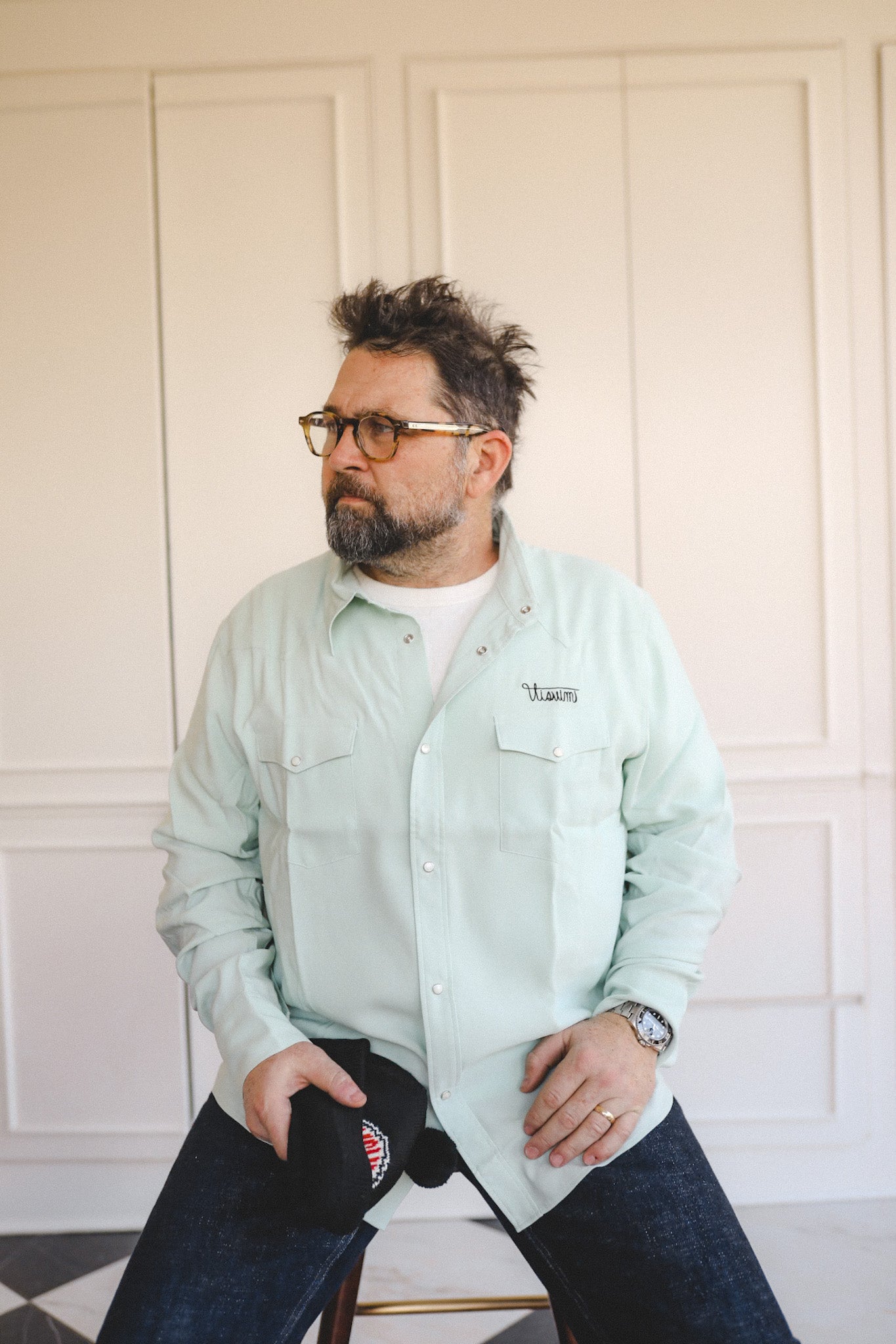Man wearing a light green shirt with a logo, sitting on a chair against a white paneled wall.