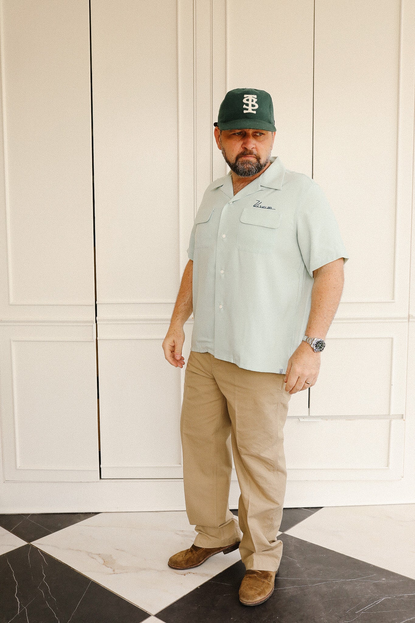 Man wearing a light green shirt, beige pants, and a cap indoors.