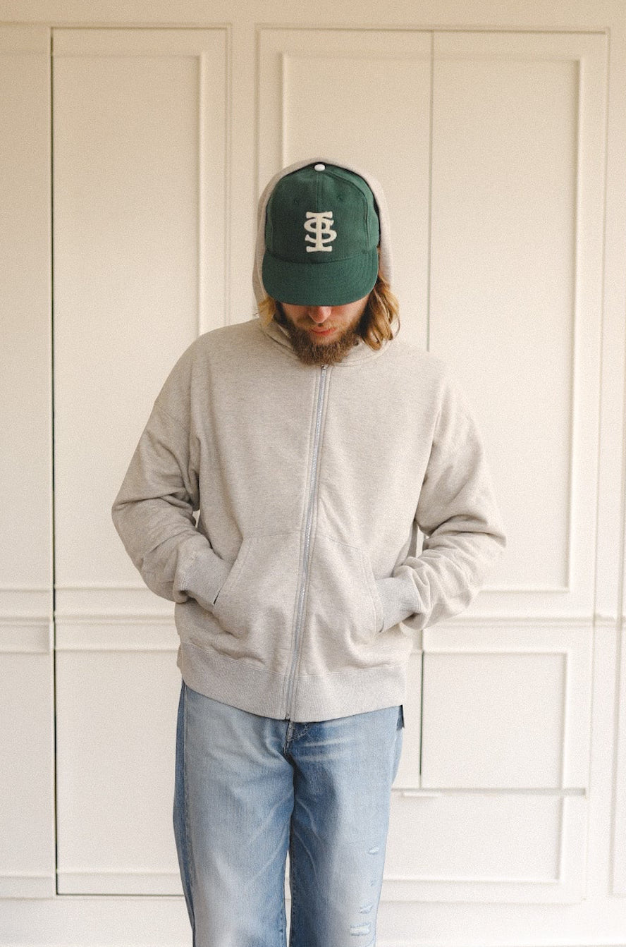 Person wearing a gray sweatshirt, blue jeans, and a green cap standing on a patterned rug with a white paneled wall in the background.