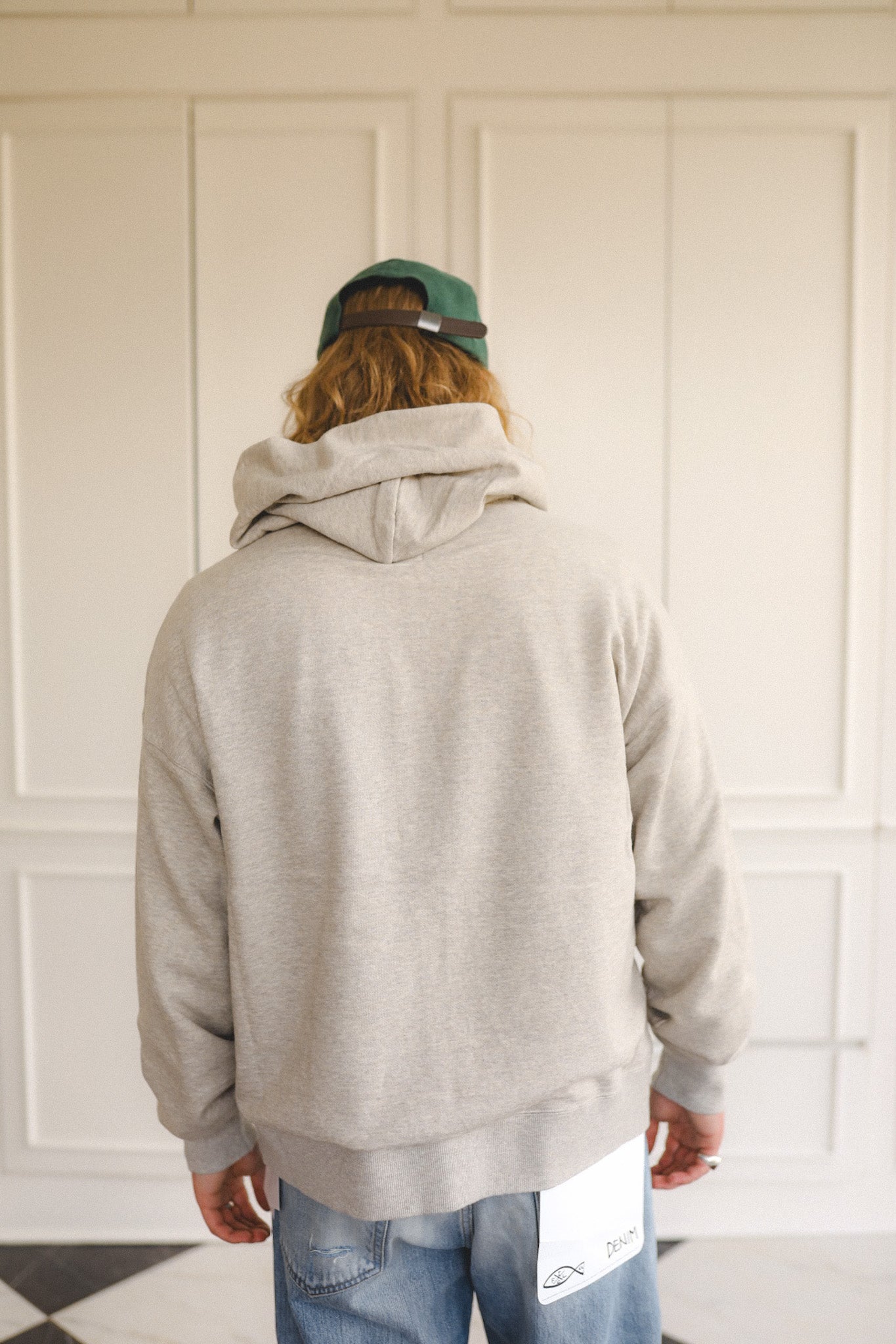 Person wearing a gray hoodie and blue jeans with a green cap, standing in a room with white paneled walls.